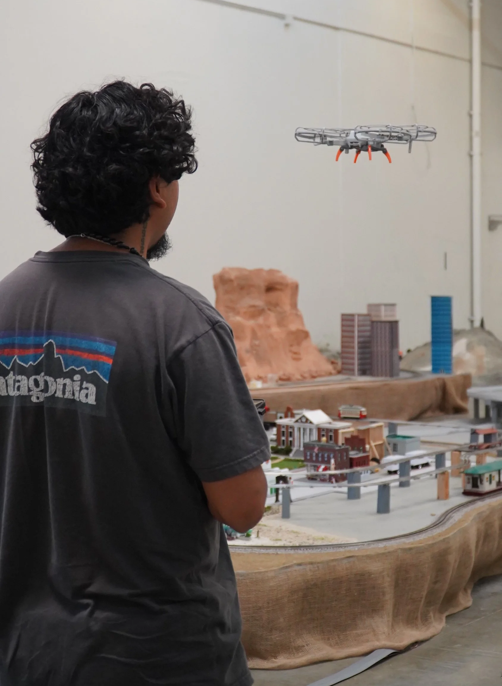 Drone student pilot flying a drone over a model train layout with miniature buildings and a rocky landscape in an indoor setting at California Drone Academy.
