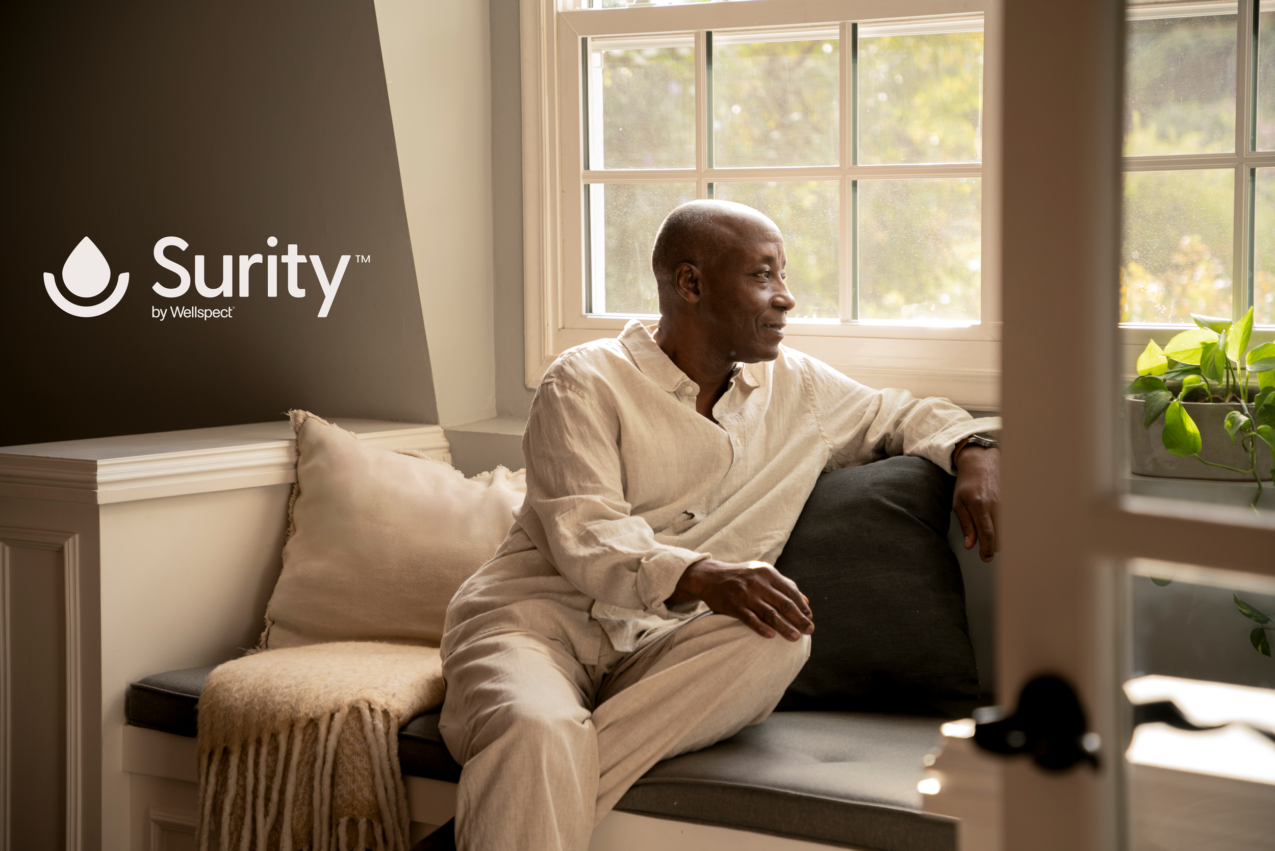 An elderly man with a bald head and dark skin sitting on a window seat inside a home. He is wearing a light-colored, long-sleeved shirt and light-colored pants, looking content. There is a window behind him with a view of trees outside, and a plant with green leaves on the right side of the window. The sofa has a pillow and a blanket on it, and a dark side table is partially visible below the window.