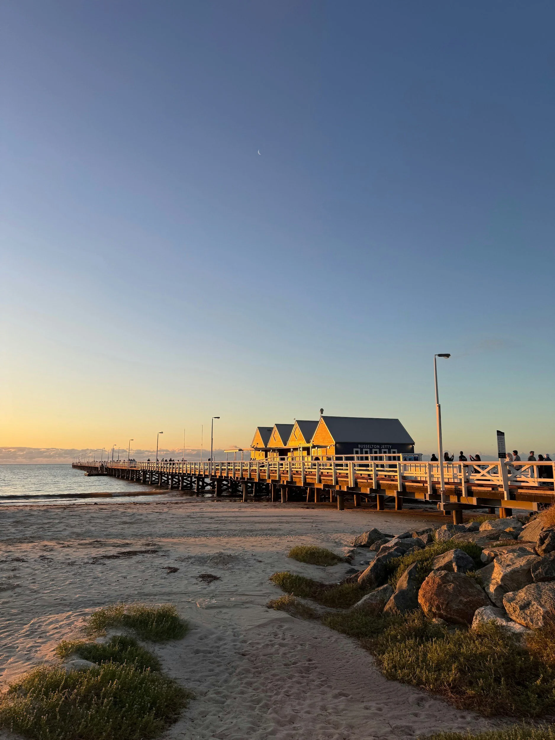 Sunset at the Busselton Jetty, rocky shoreline on the right, people walking and sunset sky with a crescent moon.