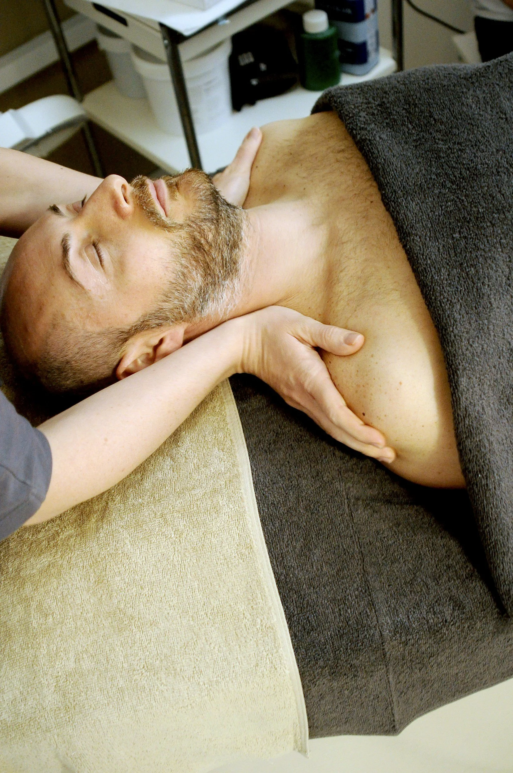 A man receiving a massage, lying down with eyes closed, partially covered with a towel.