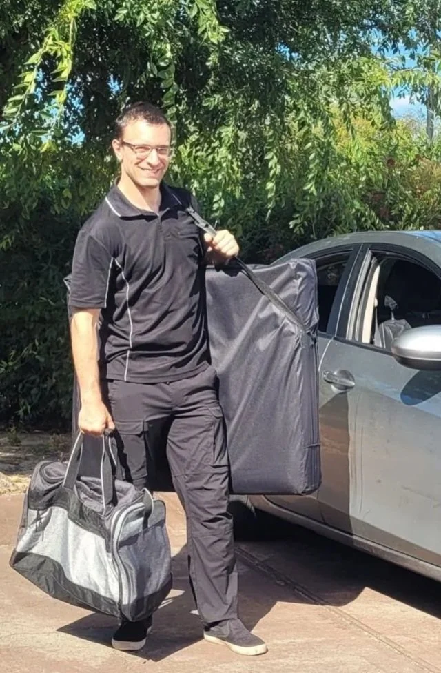 A male massage therapist standing outside next to a gray car, holding a gray and black sports bag in one hand and a packed massage table in the other, with green trees in the background.
