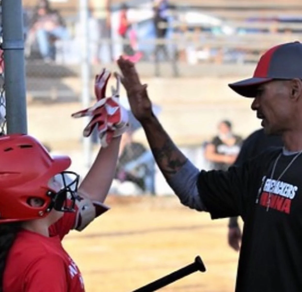 A young baseball player in a red helmet and uniform giving a high five to a man in a black shirt and cap at a baseball field.