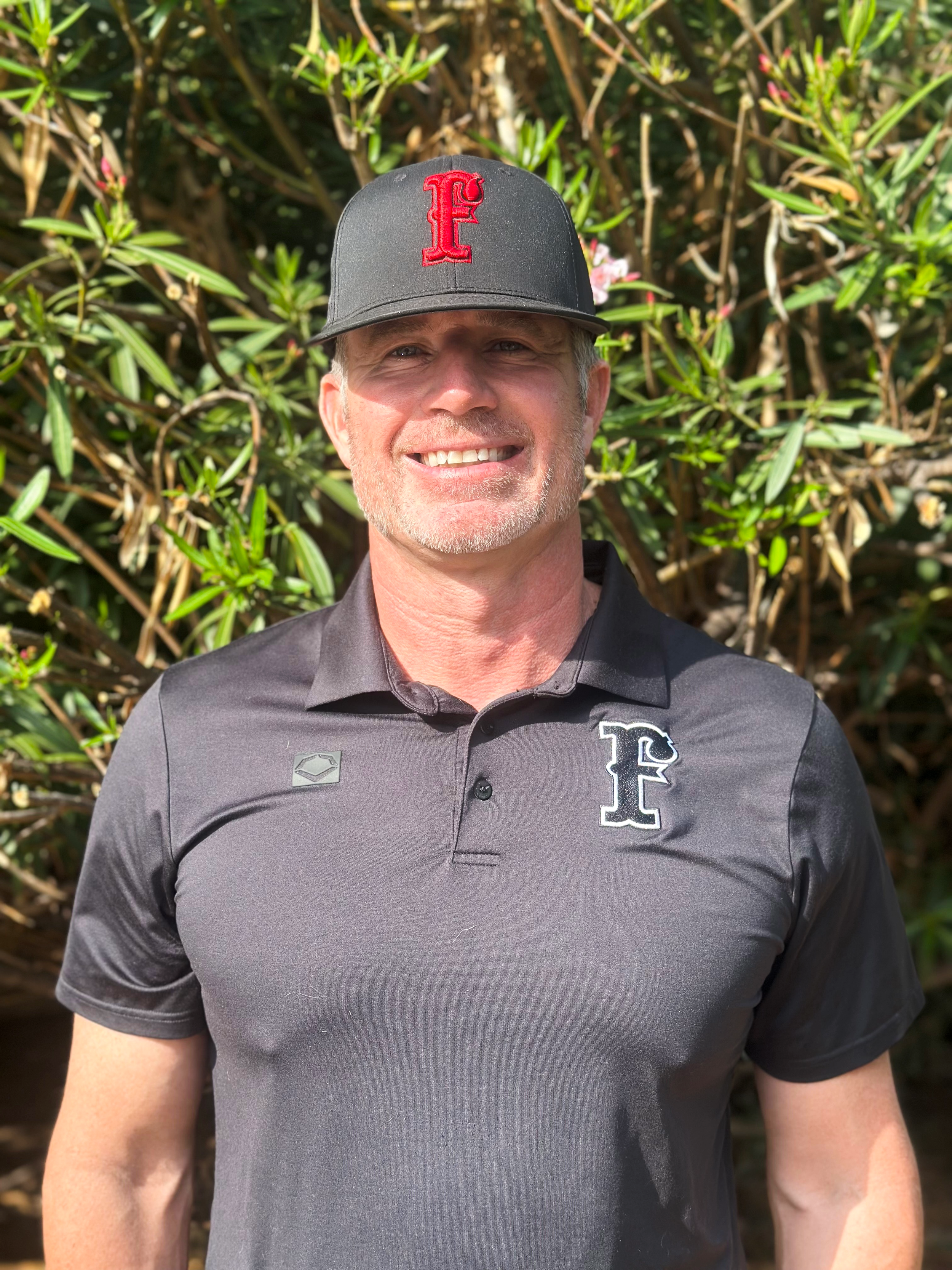 A man wearing a black baseball cap with a red 'F' logo, a black collared shirt with an 'F' logo on the chest, standing in front of green shrubbery, smiling at the camera.