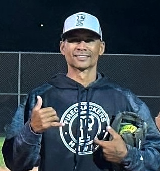 A man in a baseball cap and hoodie smiling and giving a thumbs-up at night on a sports field.