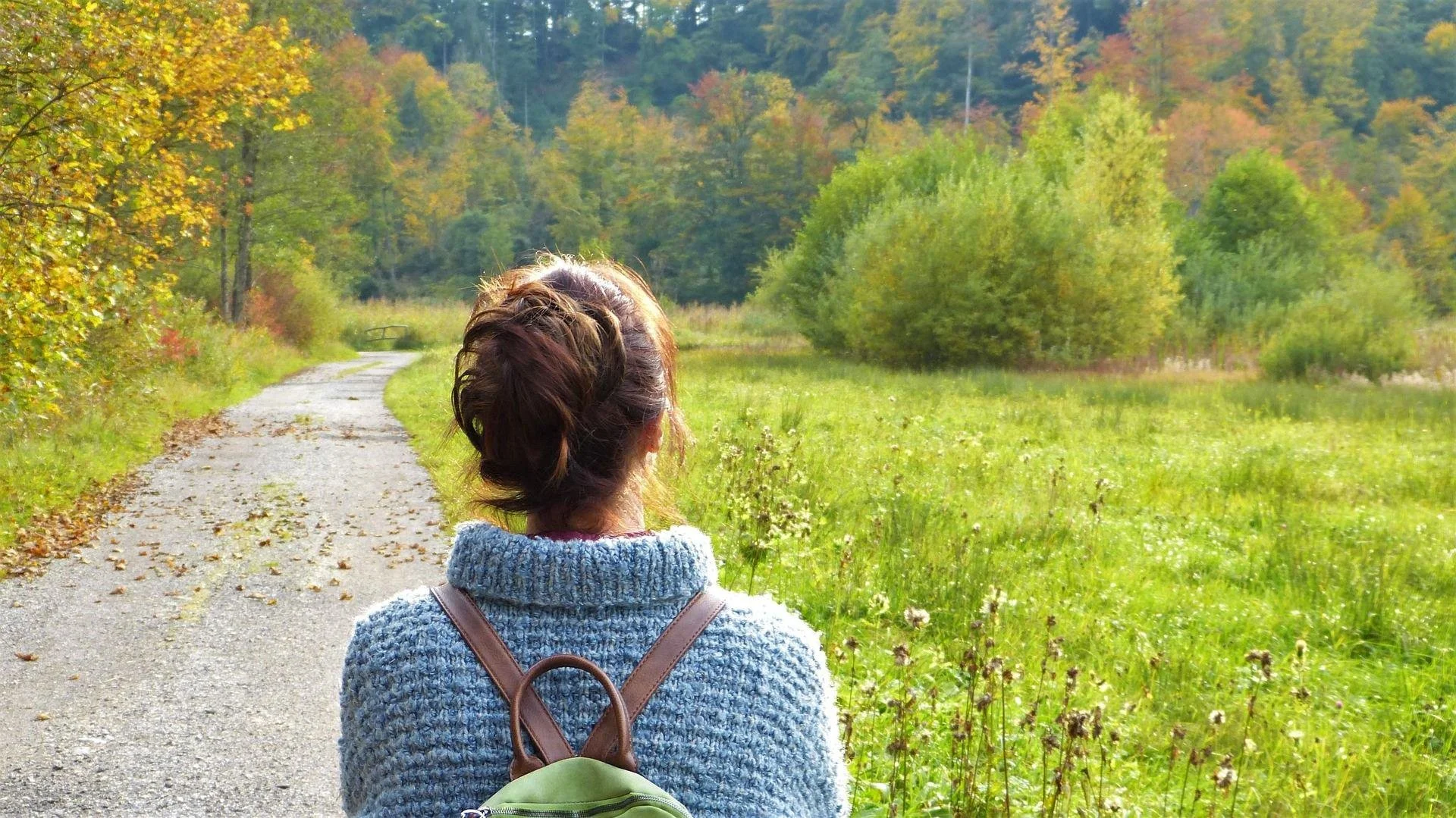 Eine Frau mit braunem, lockigem Haar trägt einen grauen Pullover und einen grünen Rucksack, während sie auf einem schmalen Weg inmitten einer grünen Wiese und Bäume mit herbstlichen Farben läuft.