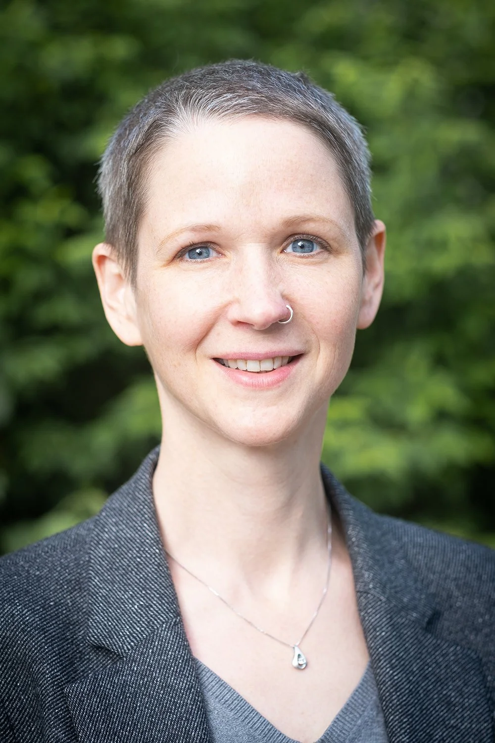 Nahaufnahme einer Frau mit kurzen, grauen Haaren, blauen Augen, einem Nasenring, lächelnd, vor grüner Natur