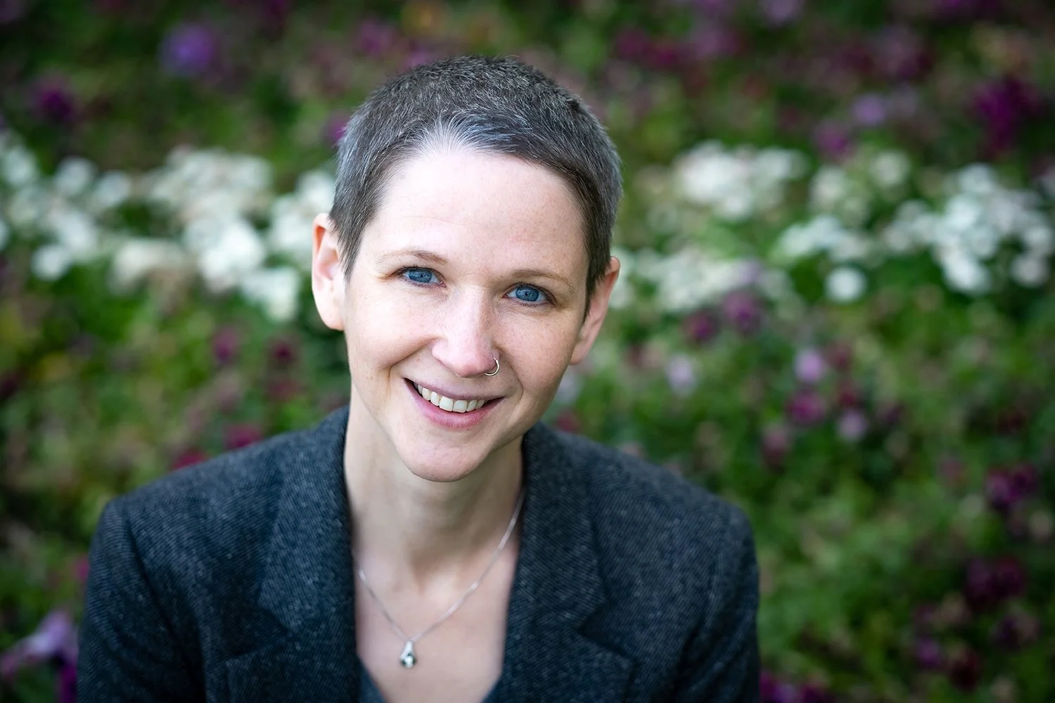 Junge Frau mit kurzen Haaren, blaue Augen, Piercing in der Nase, trägt dunkle Jacke und Kette, lächelnd vor Blumen im Garten.