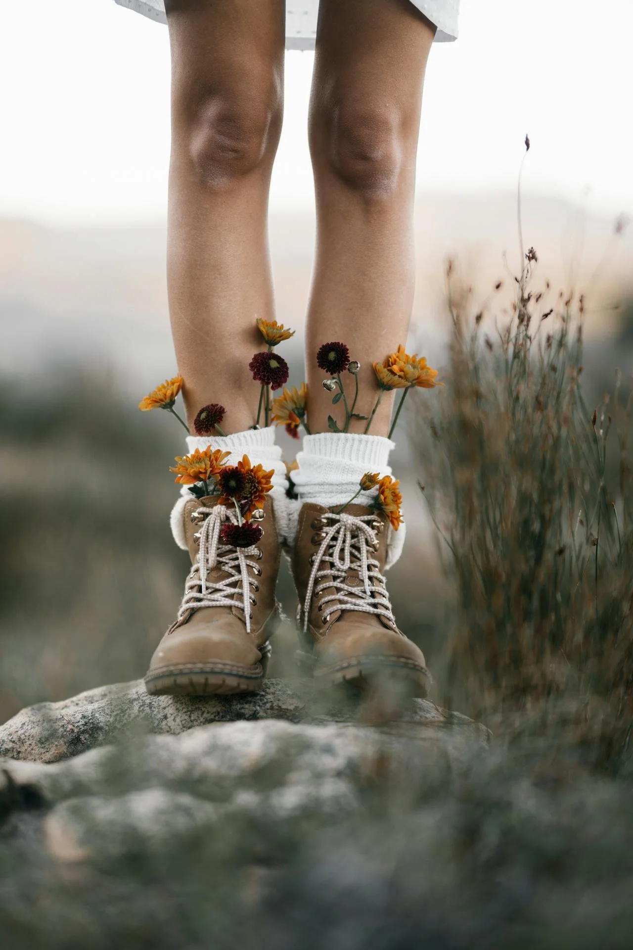 Nahaufnahme von braunen Wanderschuhen mit weißen Socken, mit bunten Blumen darin, auf einem Felsen im Freien, umgeben von Wiese und Gräsern.