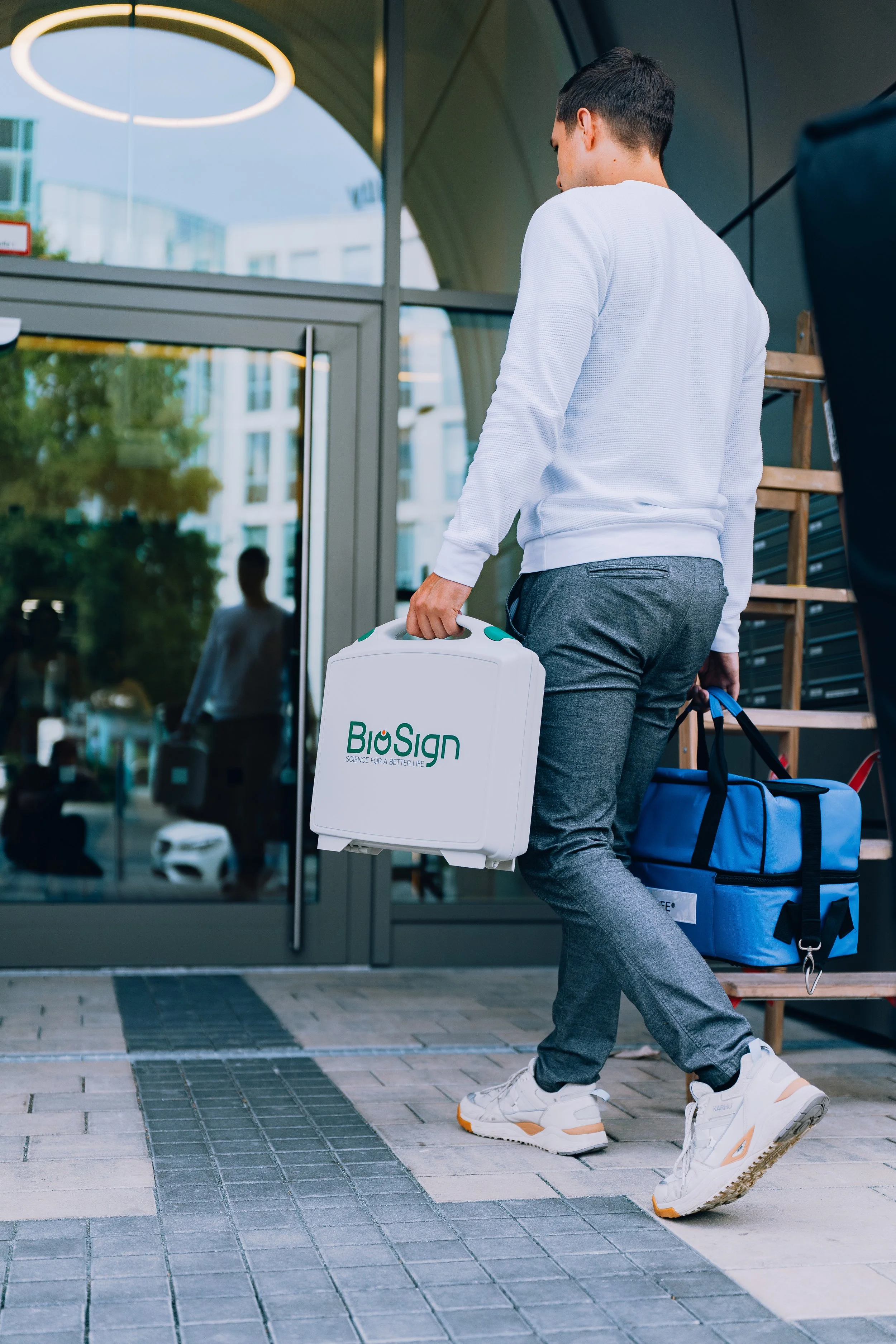 A man walking out of a building carrying a white medical cooler labeled BioSign and a blue medical bag.