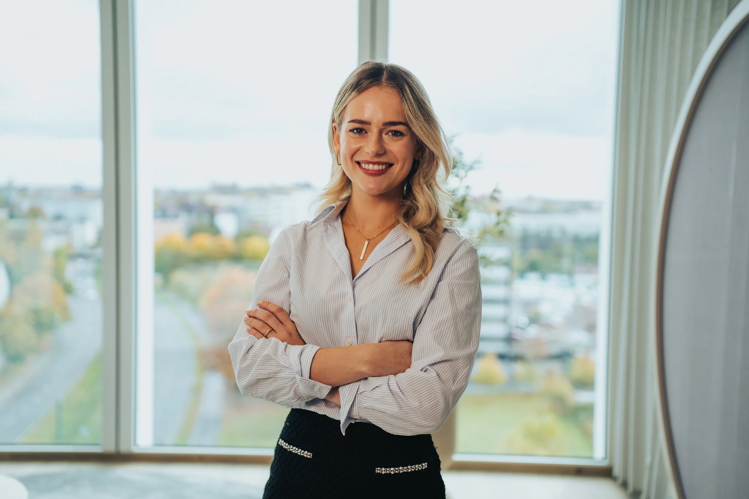 A young woman with blonde, wavy hair smiling, standing with crossed arms in front of large windows with a city landscape in the background.