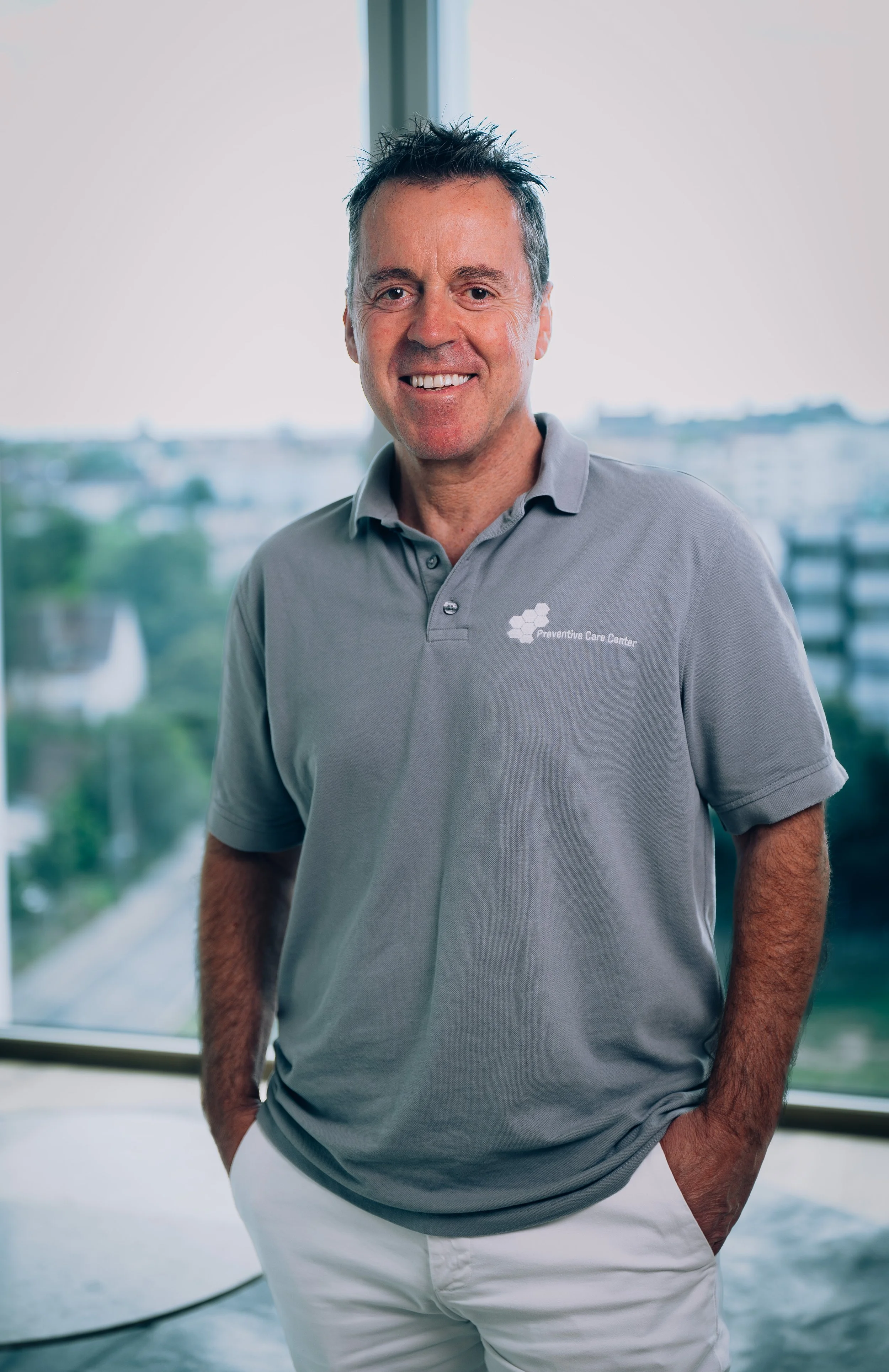 Smiling man in a white polo shirt with a Preventive Care Center logo, standing against a light, plain background.