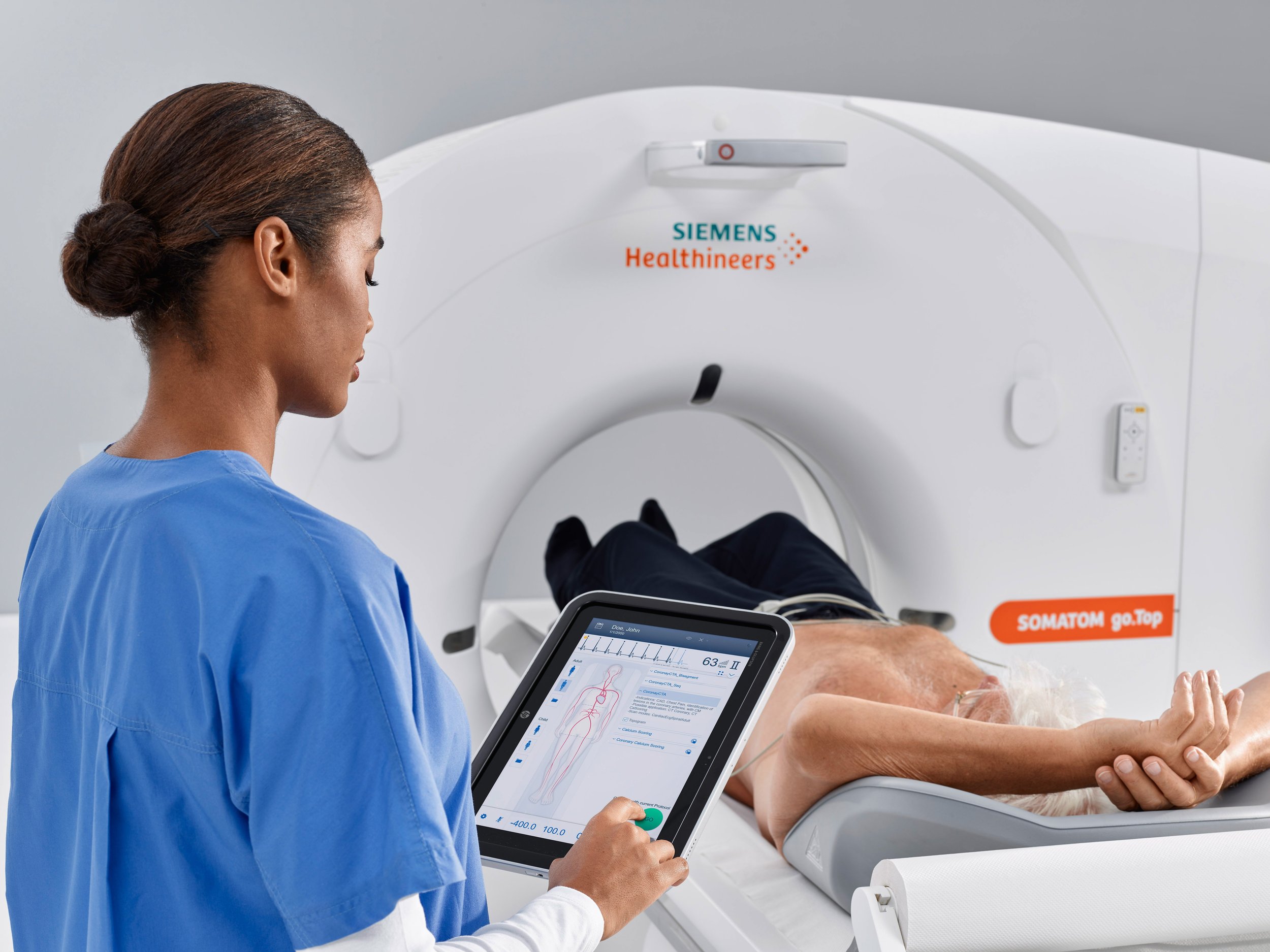 A healthcare professional in blue scrubs operating a medical imaging machine with a patient lying inside. The professional holds a tablet displaying a digital diagram of the patient's body.