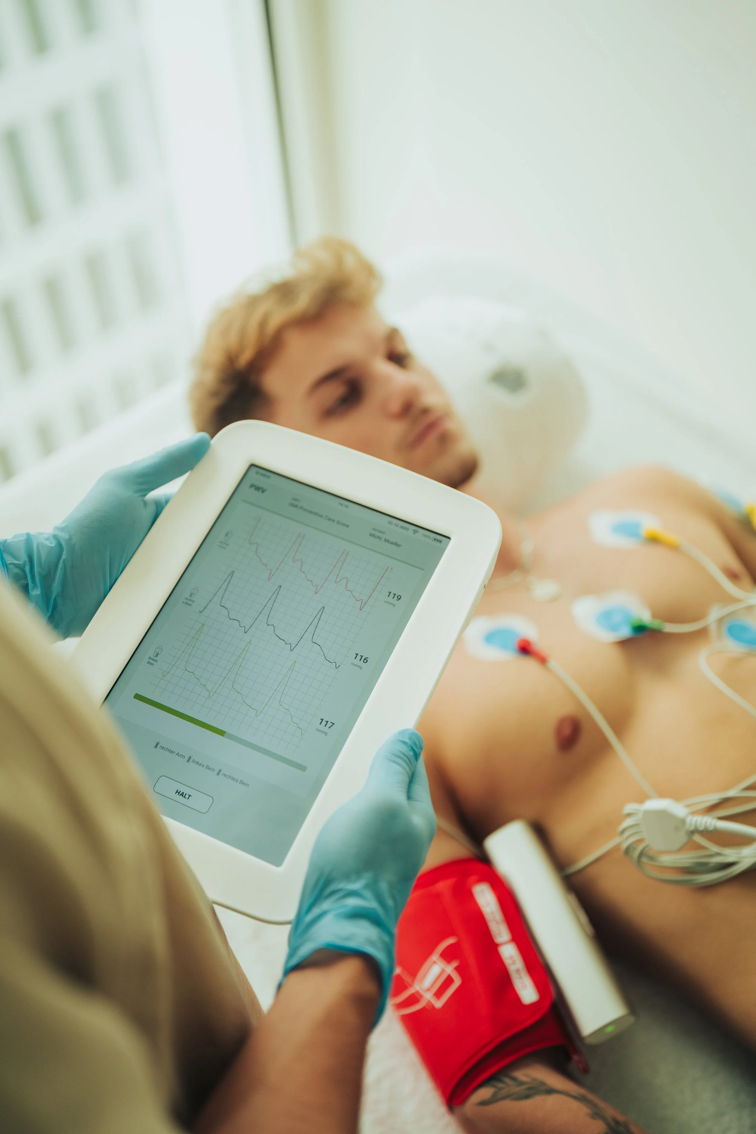 A male patient lying in a hospital bed connected to medical monitoring equipment, with a healthcare professional checking an ECG device displaying heart rate readings.