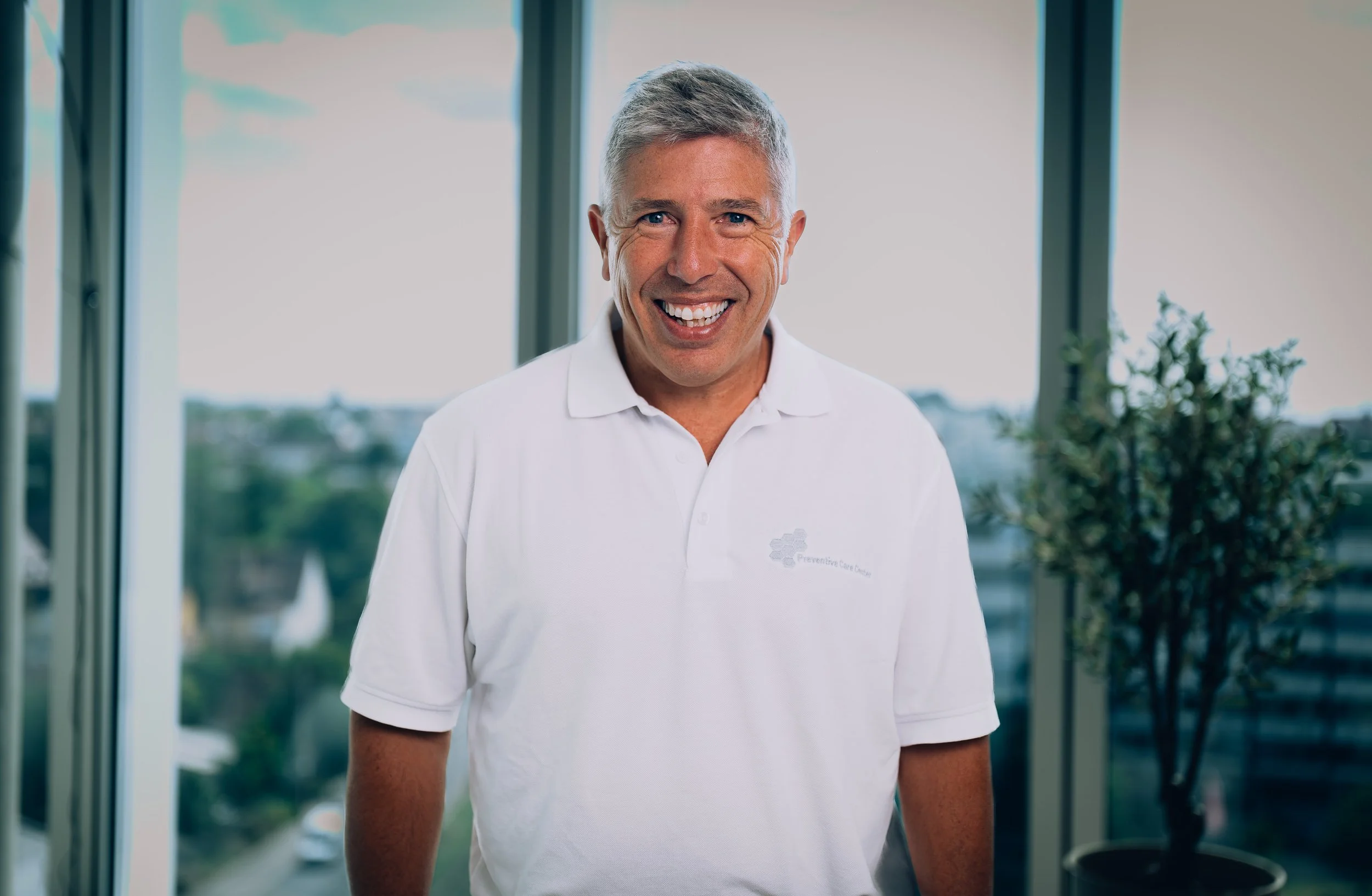 A middle-aged man with short gray hair, blue eyes, and a friendly smile, wearing a white coat and a blue tie, standing in a medical or clinical environment.