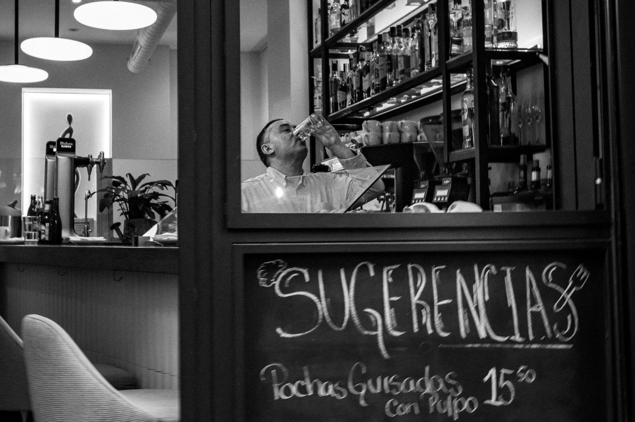 Un hombre sentado dentro de un establecimiento de alimentos bebiendo de un vaso, visto a través de una ventana de vidrio con cartel que dice 'Sugerencias' y otras palabras en español.