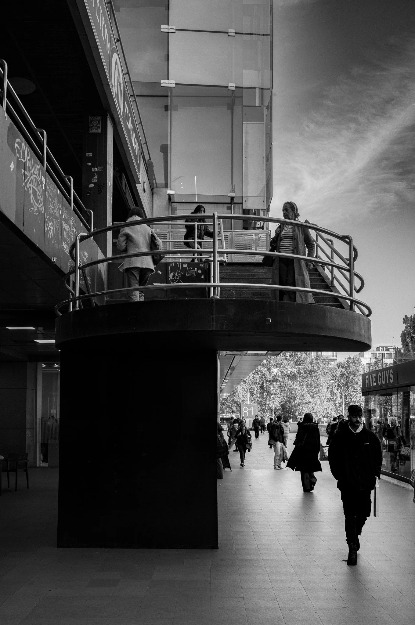 Escalera exterior en un centro comercial con varias personas caminando y tocando la barandilla, en blanco y negro.