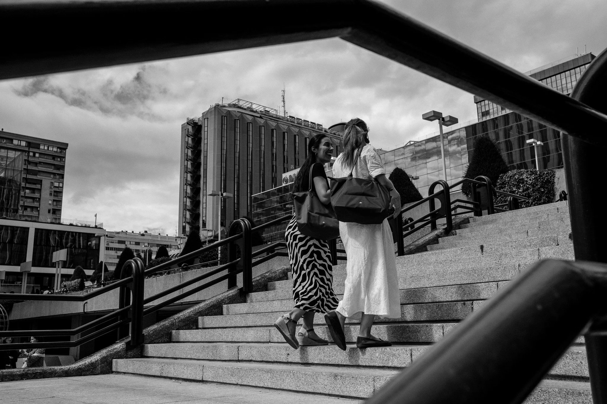 Dos mujeres caminando por unas escaleras en una zona urbana, vista desde abajo, con edificios altos de fondo y cielo nublado.