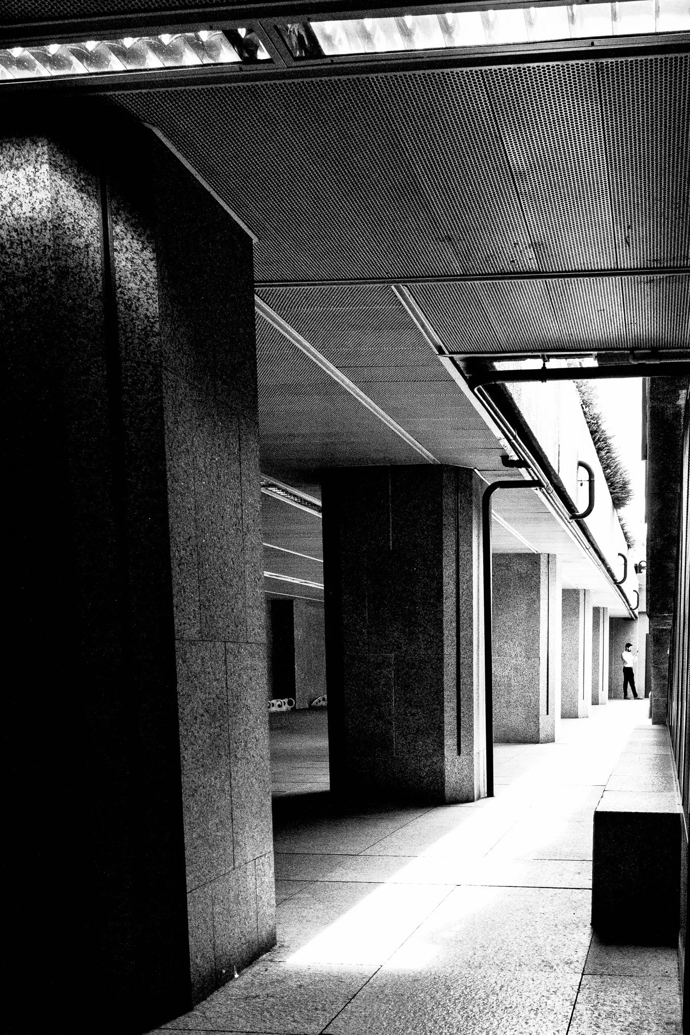 Pasillo exterior en negro y blanco con columnas de piedra, suelo de baldosas, protecciones en la orilla y un hombre de pie en el fondo.