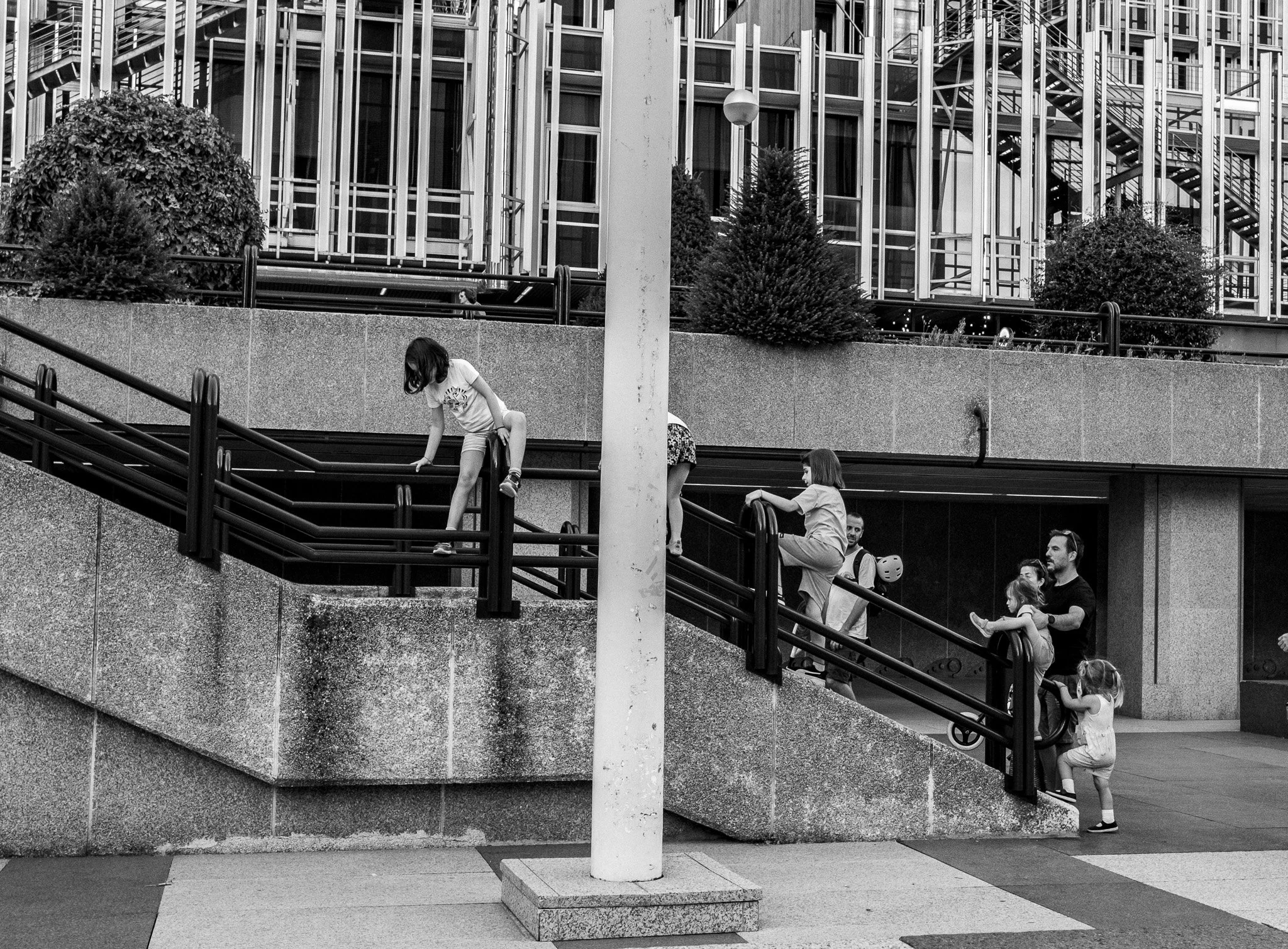 Niños y adultos en una zona de juegos urbana, algunos subiendo escaleras y otros en la parte superior, con edificios modernos al fondo