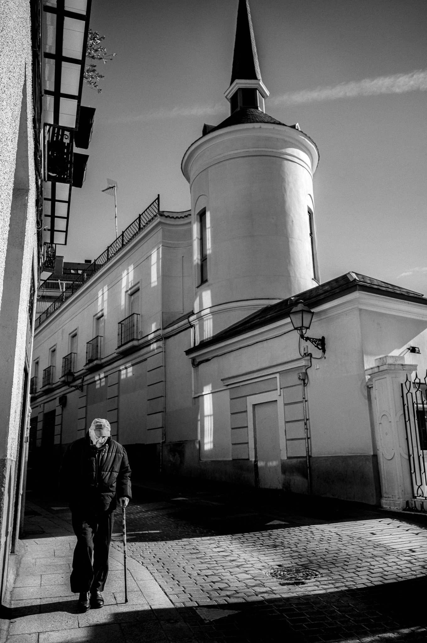 Una calle con un edificio antiguo con torre en el fondo y una persona mayor caminando con bastón en primer plano, en blanco y negro.
