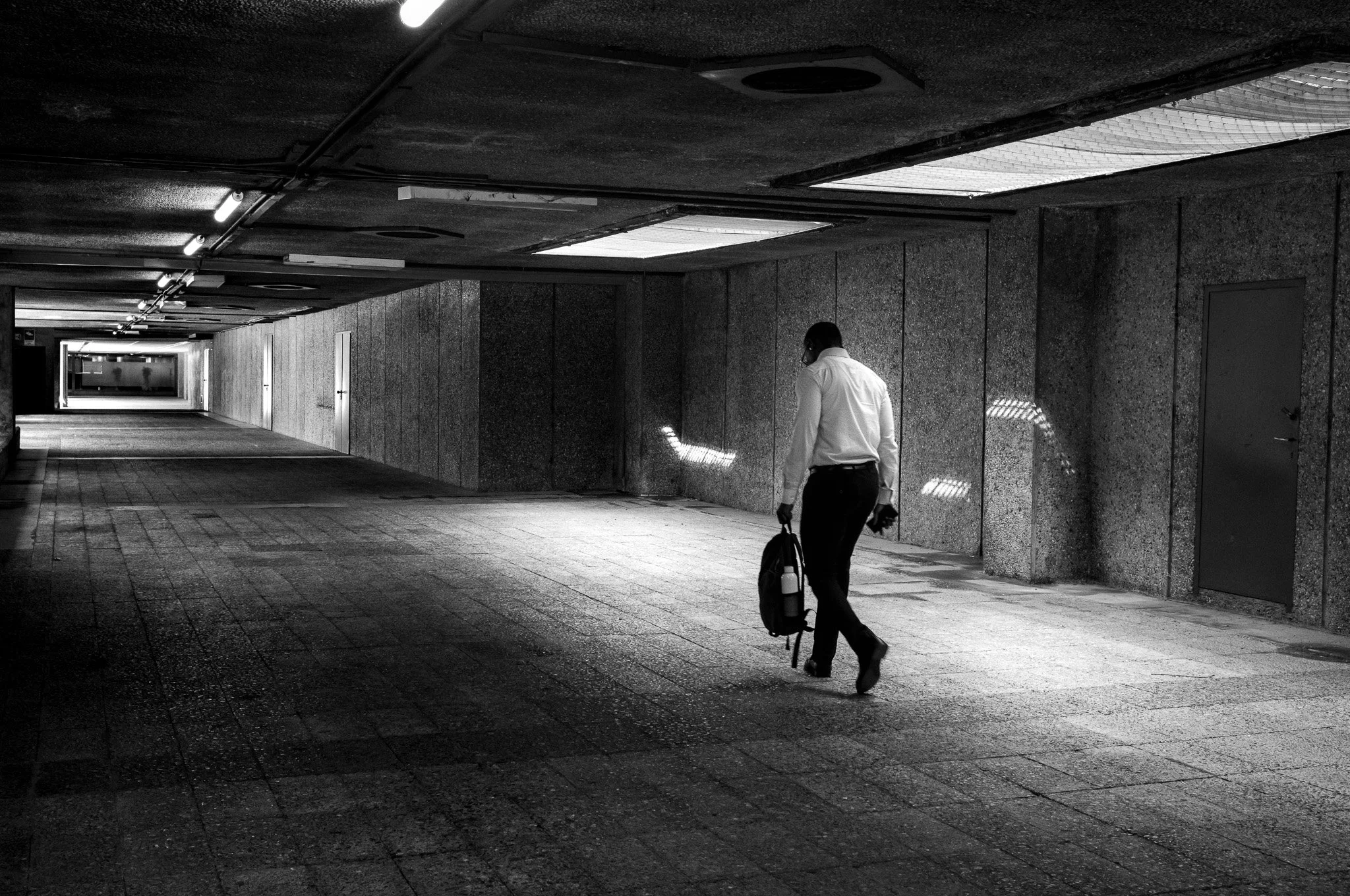 Hombre de negocios caminando por un pasillo subterráneo, vestido con camida de vestir blanco y pantalones oscuros, llevando una mochila y un teléfono móvil.