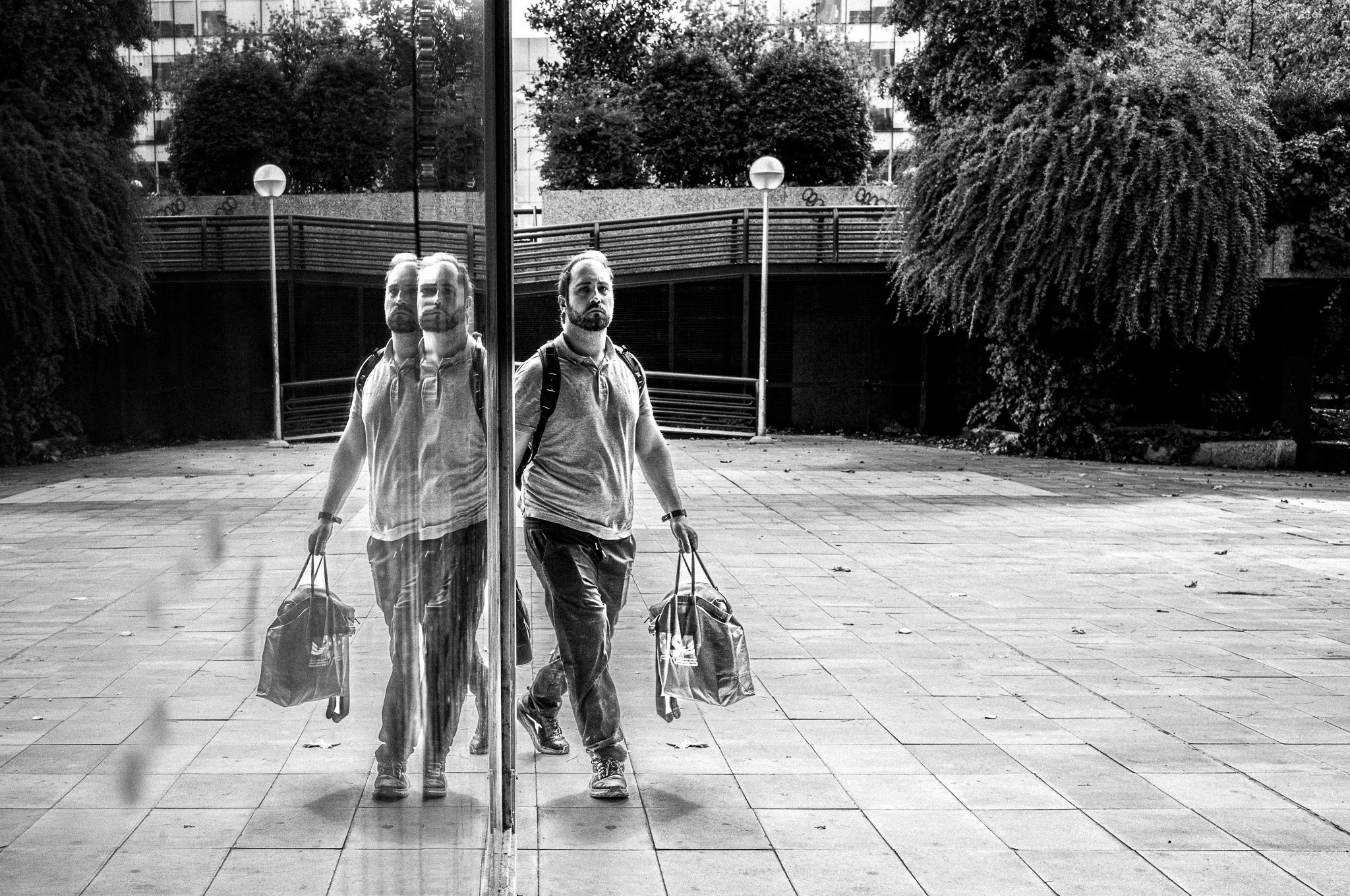 Hombre con mochila y bolso de mano reflejado en una estructura de cristal al aire libre.