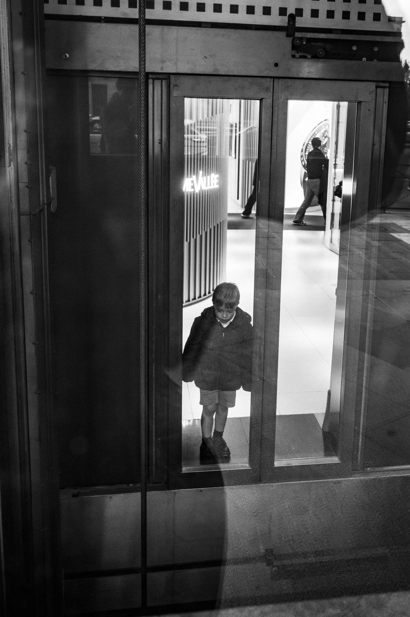 Niño mirando por la puerta de un edificio, en un entorno urbano, en negro y blanco.