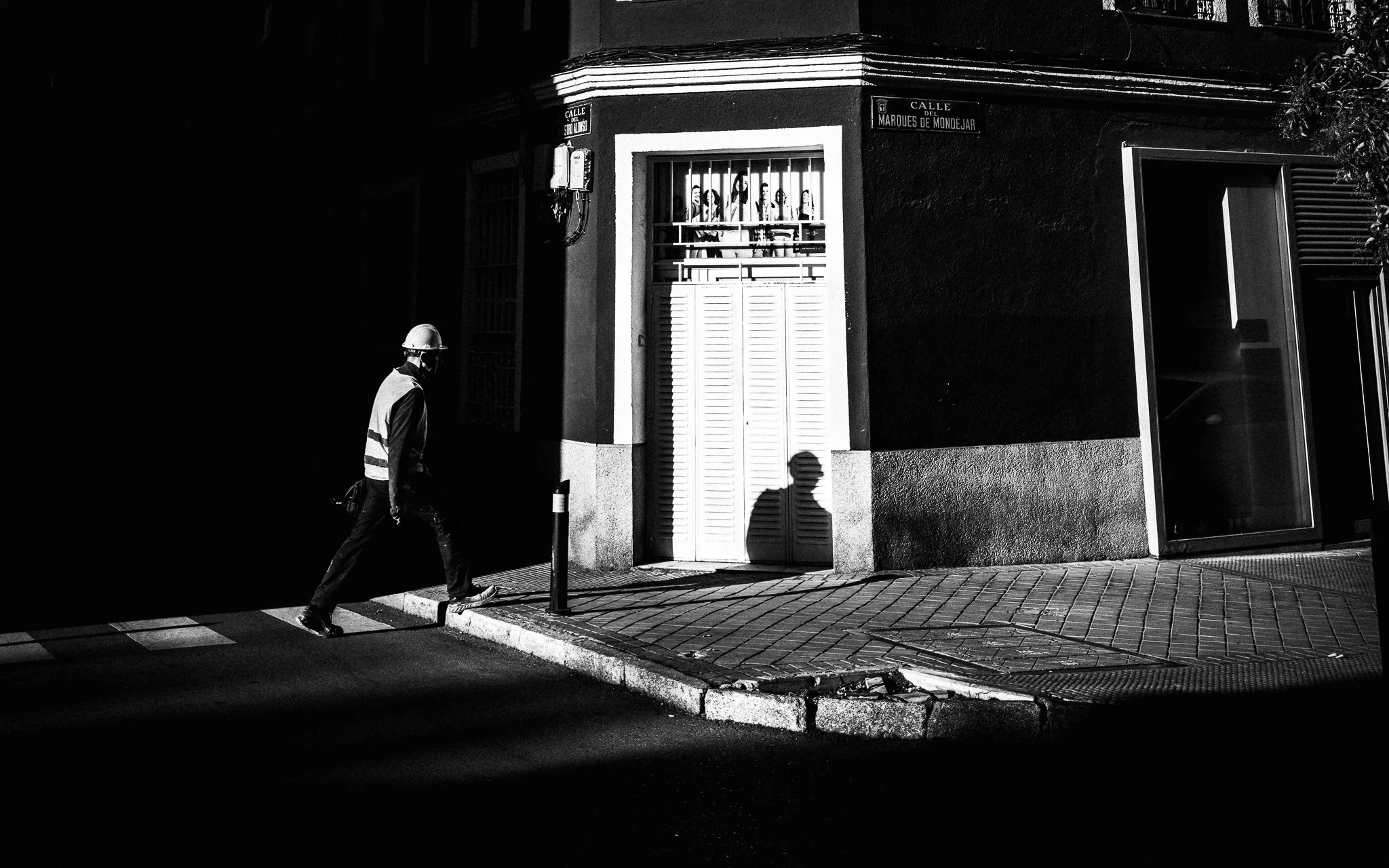 Una calle en una ciudad de noche en blanco y negro, con un hombre con casco y chaleco reflectante caminando por la acera y la sombra de un hombre en la pared frente a la puerta.