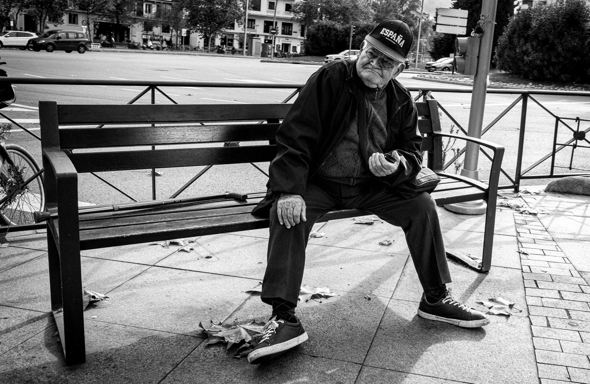Hombre mayor sentado en un banco de parque en la calle, sosteniendo un teléfono móvil, usando una gorra que dice 'España'.