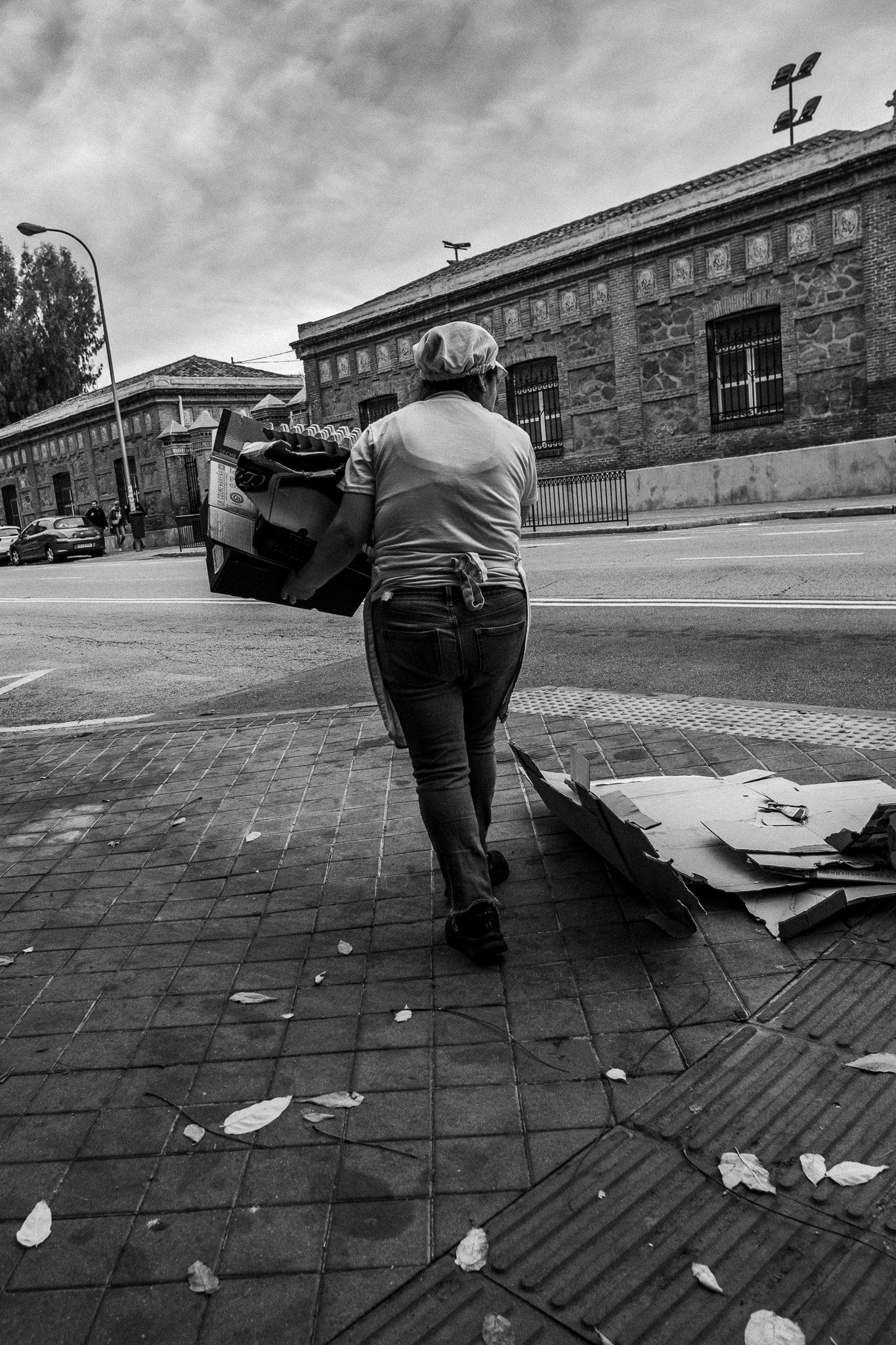 Mujer caminando con cajas en la calle, vista de espaldas, en un entorno urbano con edificios antiguos y un cielo nublado.