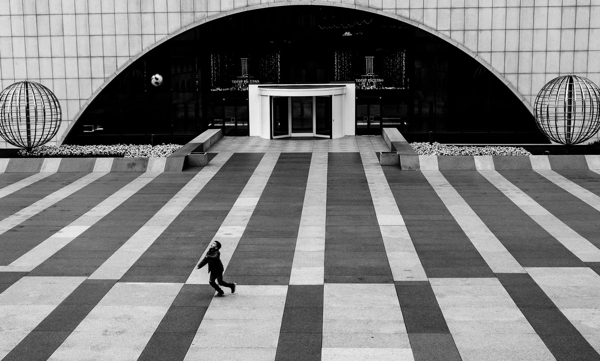 Una vista en blanco y negro de una entrada moderna a un edificio con un arco grande y columnas blancas en la parte superior, con un niño caminando sobre un cruce peatonal amplio y colorido en primer plano.