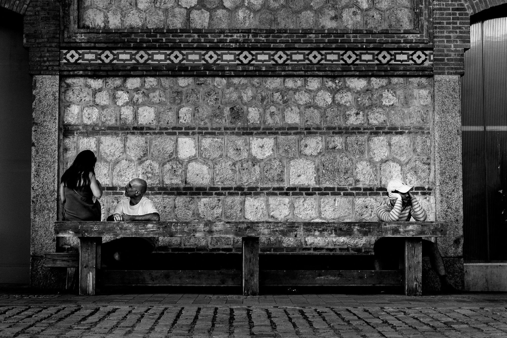Escena en la calle con dos personas conversando y una niña que tiene la cabeza agachada, todos en un banco de madera frente a un muro de piedra con detalles de azulejos.