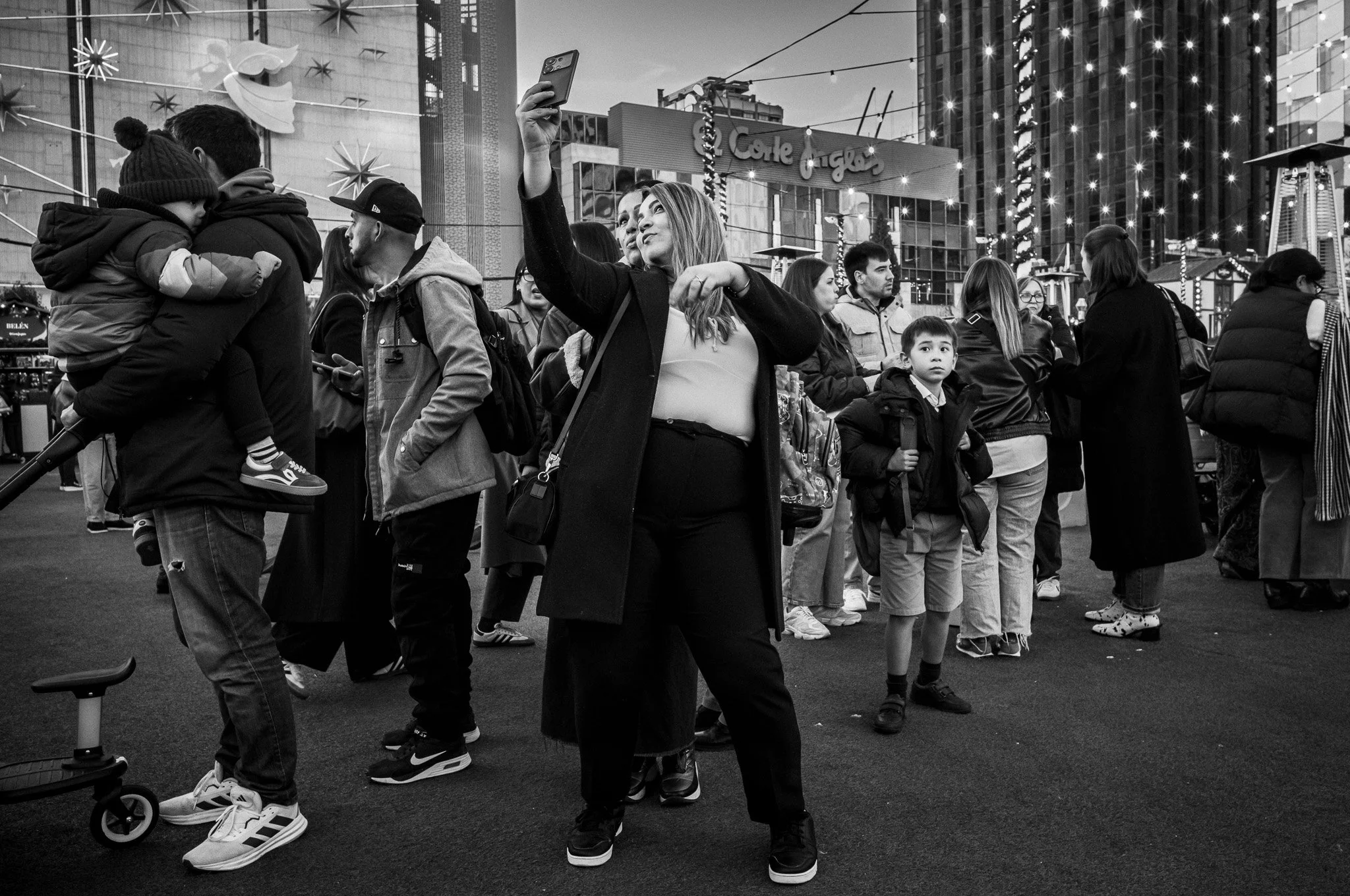 Persona tomando una selfie en una plaza concurrida en la ciudad, con grupos de personas y luces decorativas en el fondo.