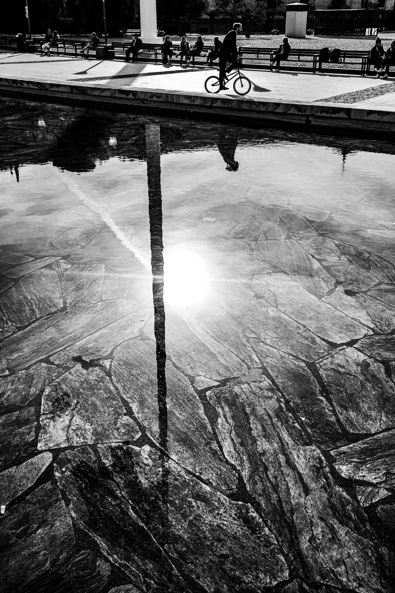 Una vista en blanco y negro de una fuente con piedras, reflejando el sol y una columna, con varias personas sentadas en bancos y una persona en bicicleta en un parque