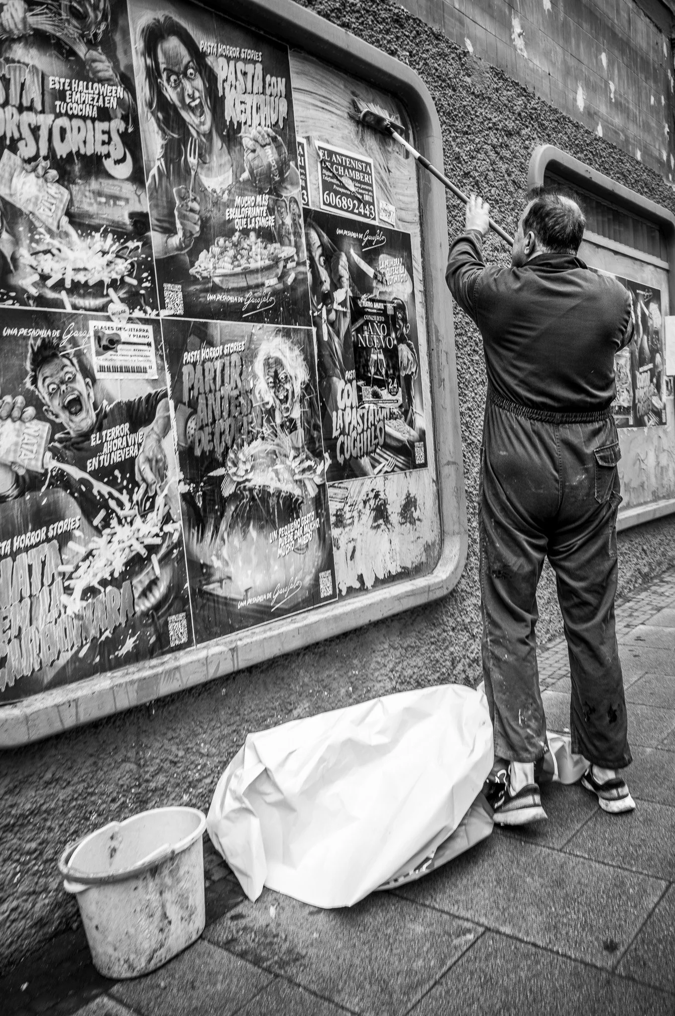 Hombre limpiando un cartel de anuncios publicitarios en la calle con una escoba y cubo de basura al lado, en un entorno urbano.