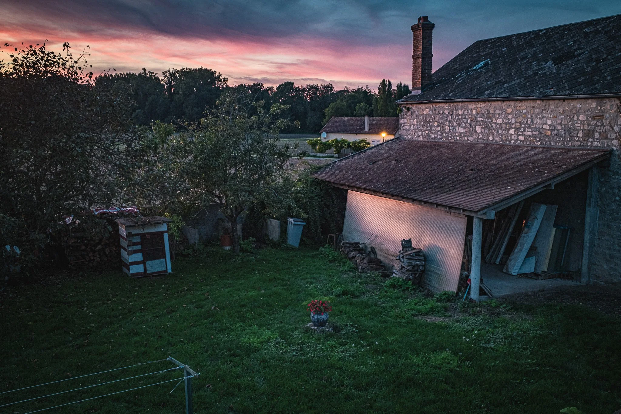 Jardín con césped, árbol, caseta para mascotas y fogón, al atardecer con cielos rosa y azul, cerca de una casa de piedra con techo de tejas y chimenea.