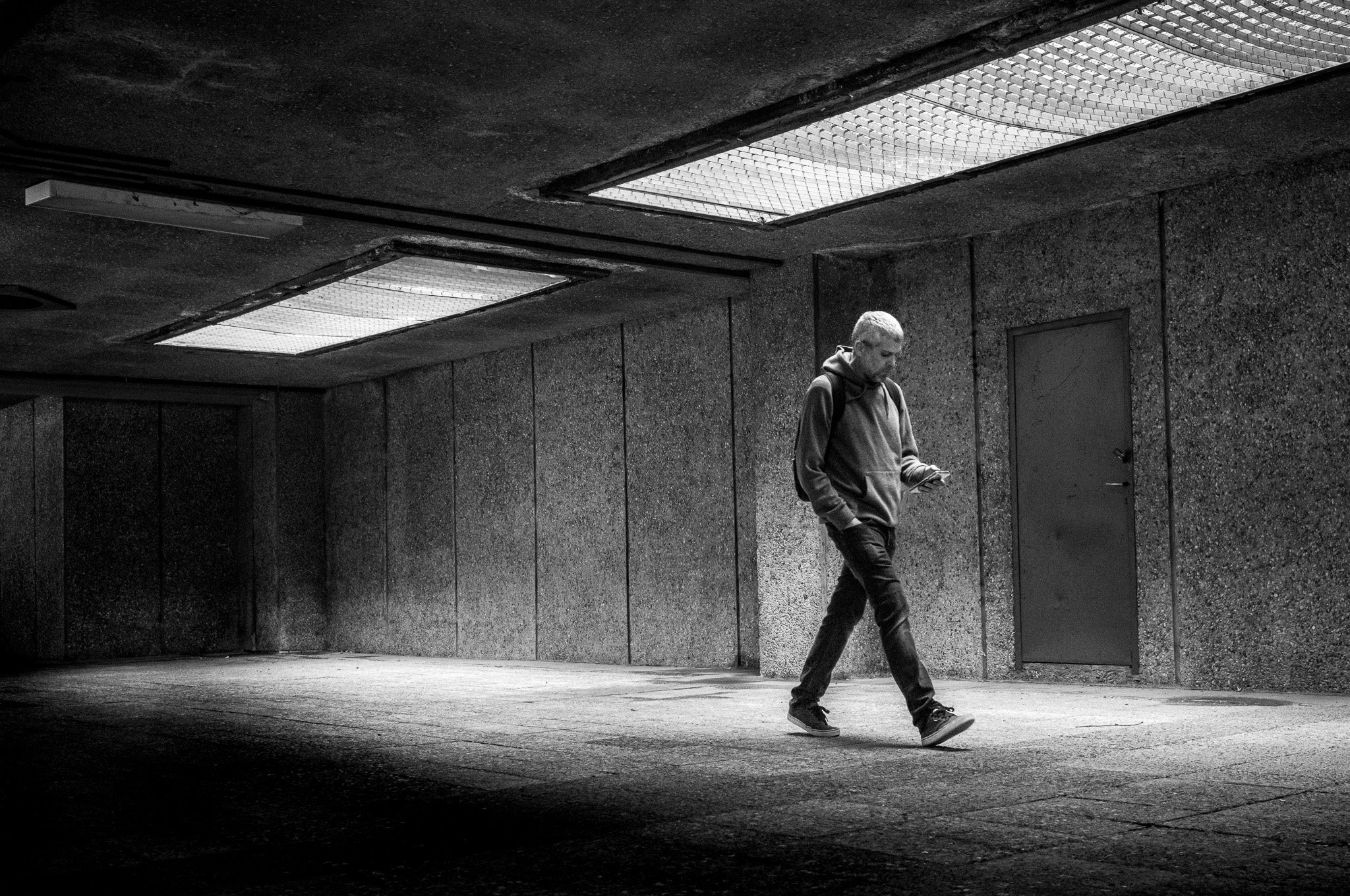 Un joven caminando solo en un paso subterráneo con paredes de concreto y luces fluorescentes en el techo, en una atmósfera en blanco y negro.
