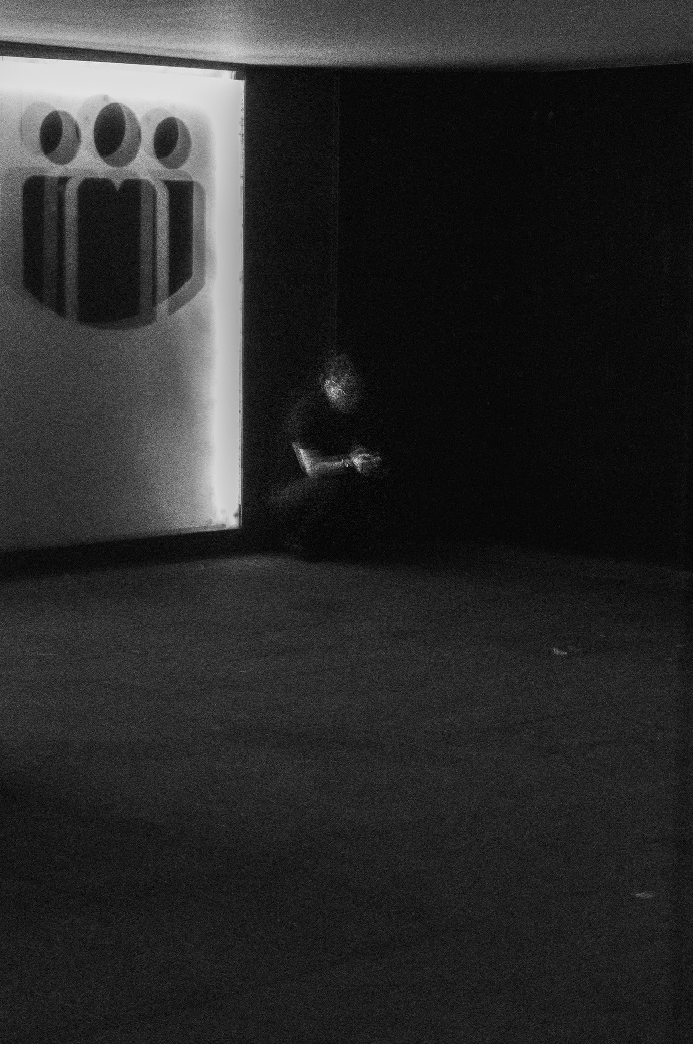 Una persona sentada en una esquina en un espacio oscuro, con un cartel brillante que tiene un símbolo de tres figuras humanas estilizadas. La persona parece estar usando un teléfono móvil.