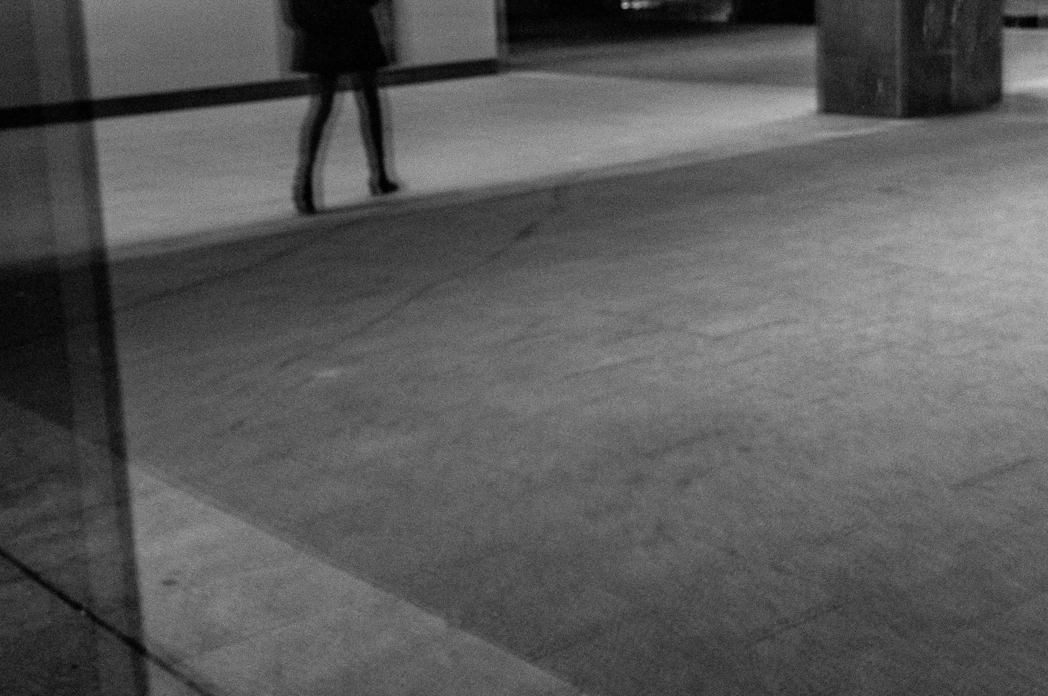 Una imagen en escala de grises que muestra un pasillo con una mesa en el fondo, una persona caminando y una columna en el lado derecho.