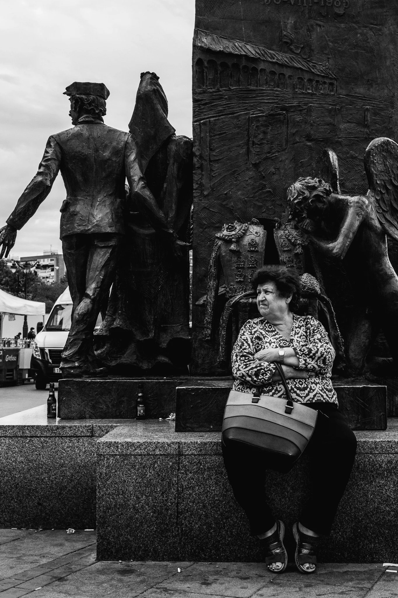 Una mujer sentada frente a una escultura de bronce en una plaza urbana, con edificios y vehículos en el fondo.