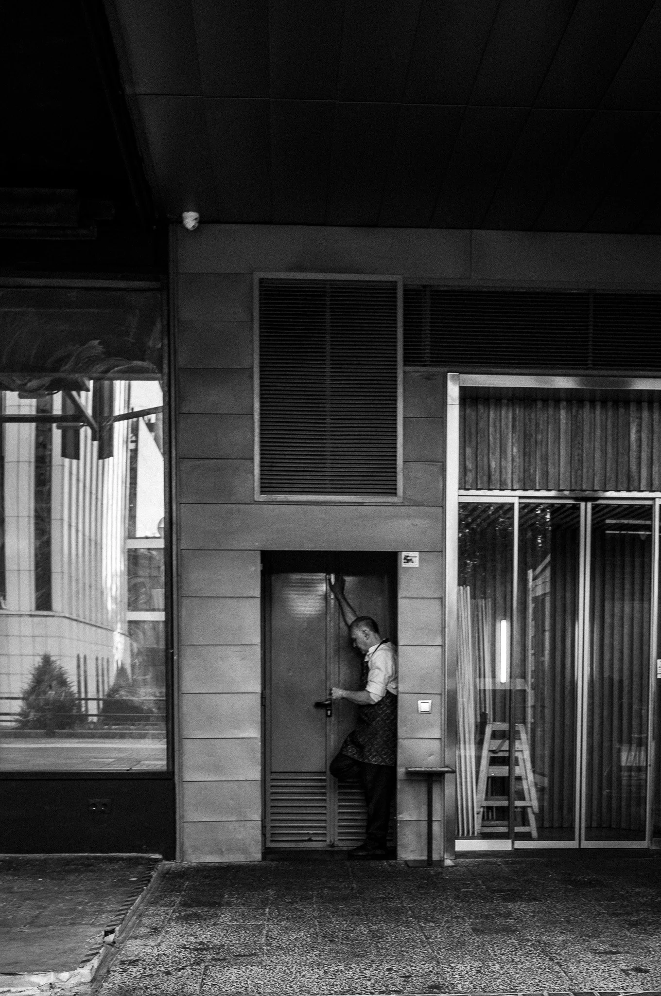Hombre de espaldas, vestido con camisa blanca y delantal, usando una linterna dentro de un edificio moderno en la ciudad, en un entorno urbano nocturno.