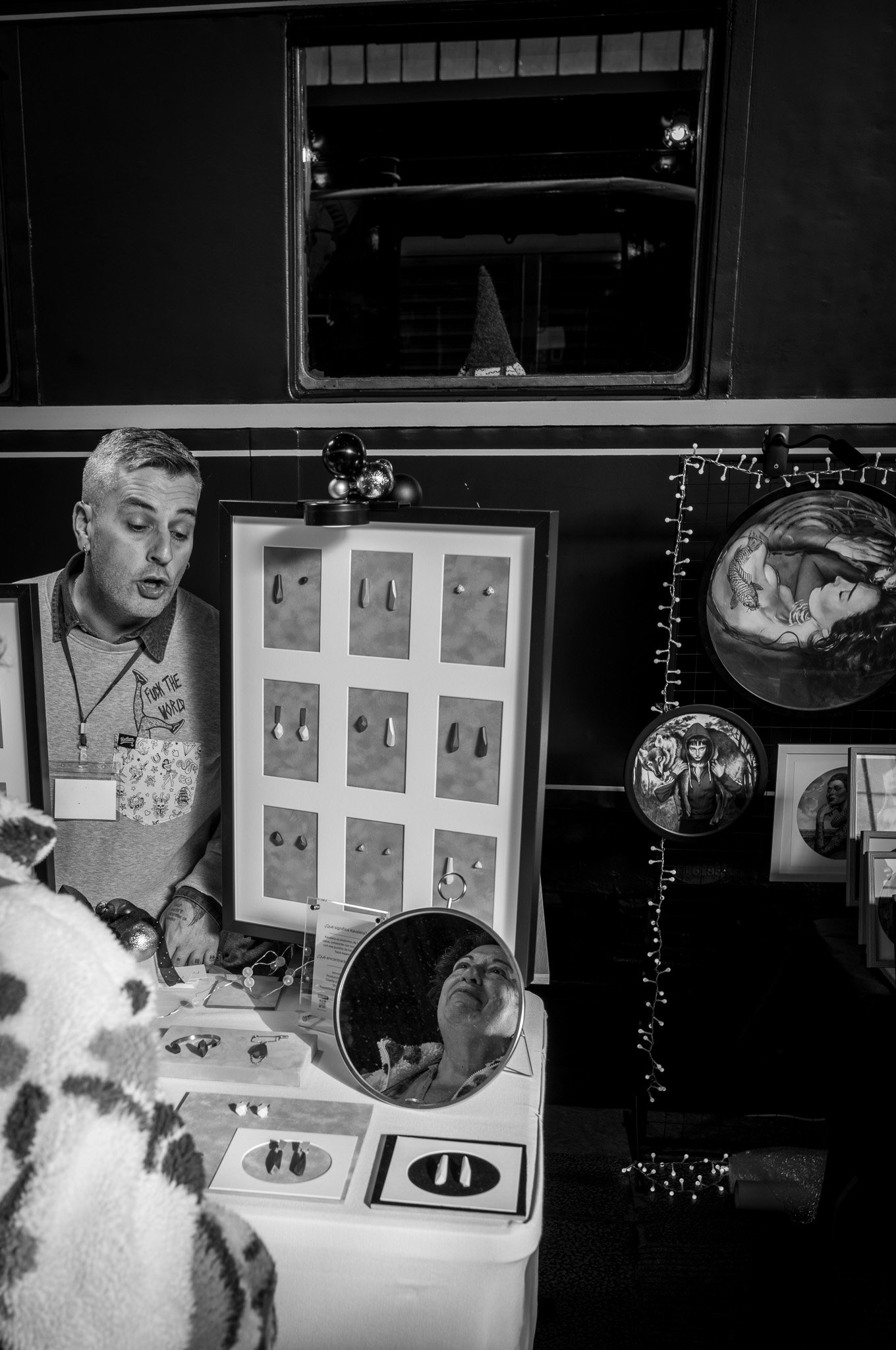 Hombre en puesto de artesanías con exhibición de pendientes, decoraciones navideñas y arte enmarcado.