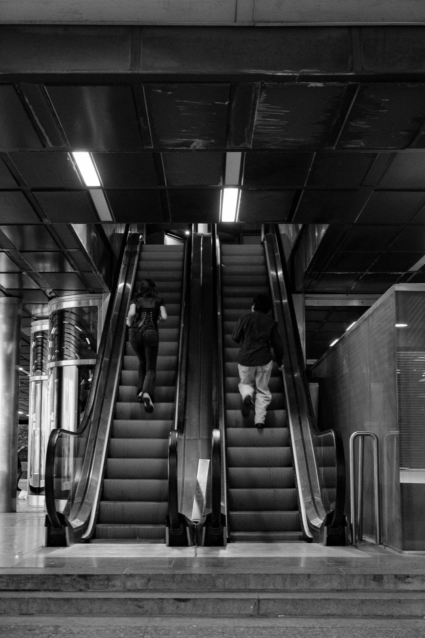 Dos personas usando las escaleras mecánicas en un espacio interior con estructura moderna y techo de paneles negros.