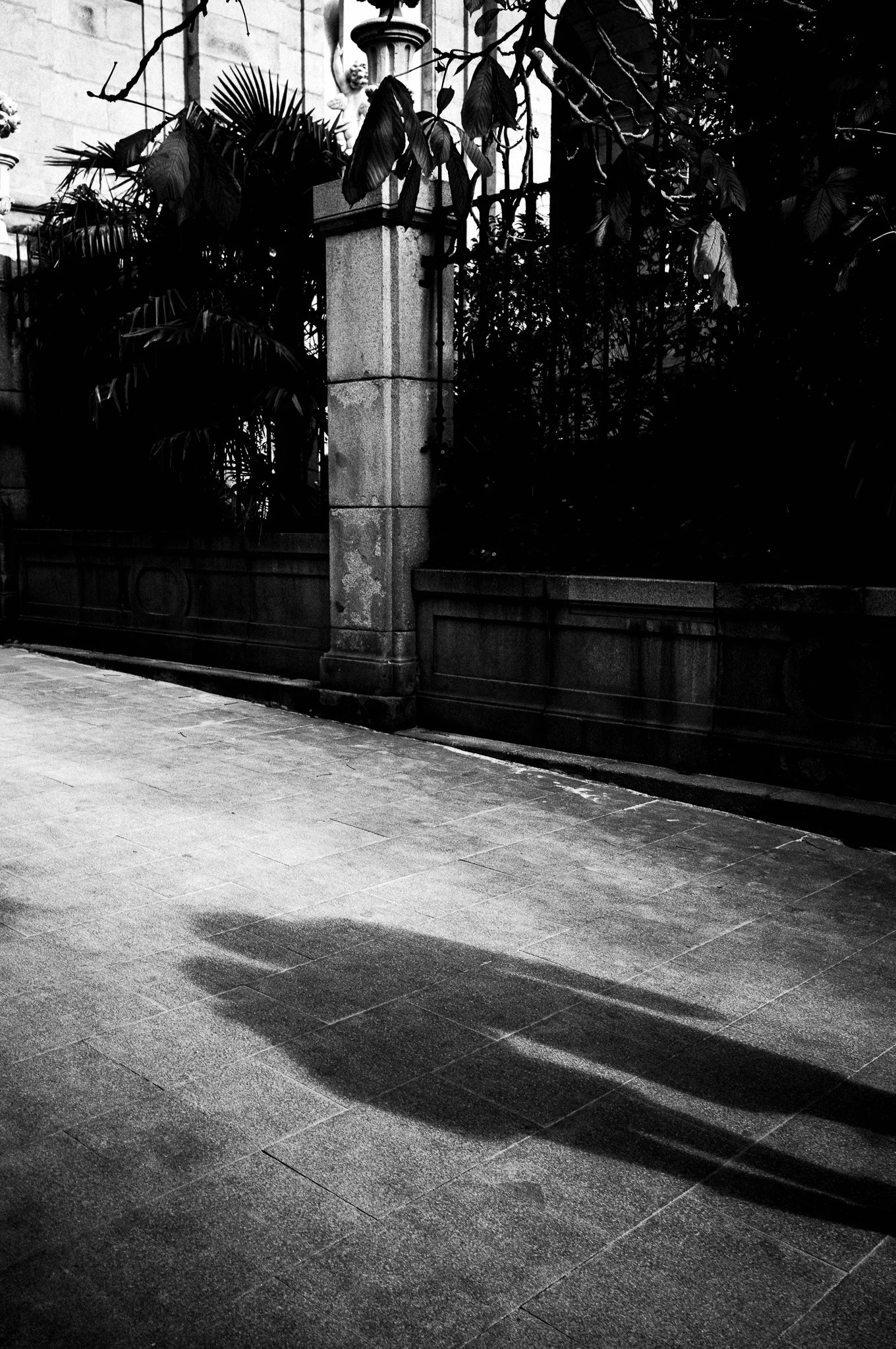 Una imagen en blanco y negro de una cerca de hierro forjado con un poste de piedra. Hay vegetación a la derecha y una estatua de un ángel en la parte superior del poste de piedra.