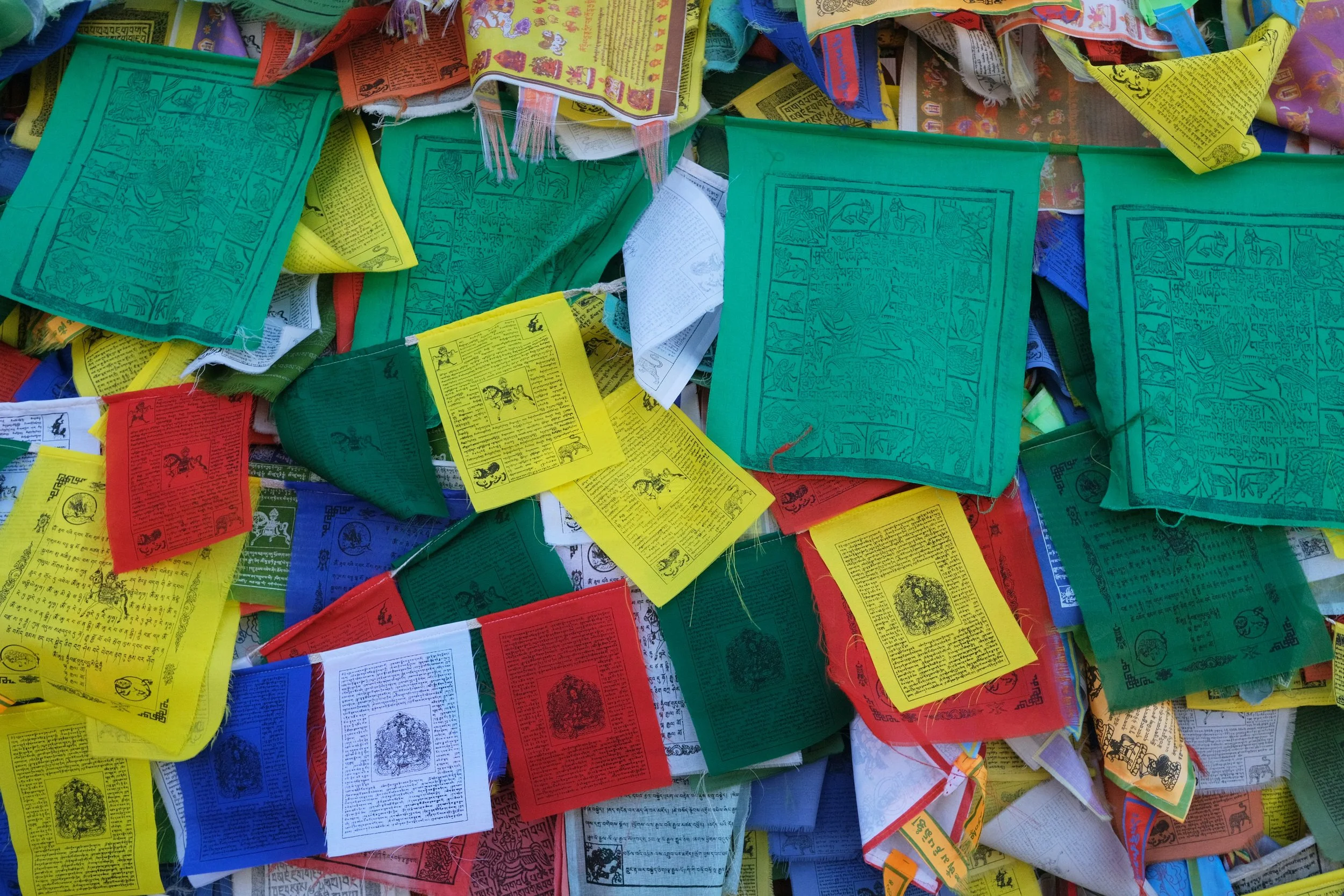 A large pile of colorful prayer flags in green, yellow, red, white, and blue, with intricate writings and illustrations, scattered and overlapping.