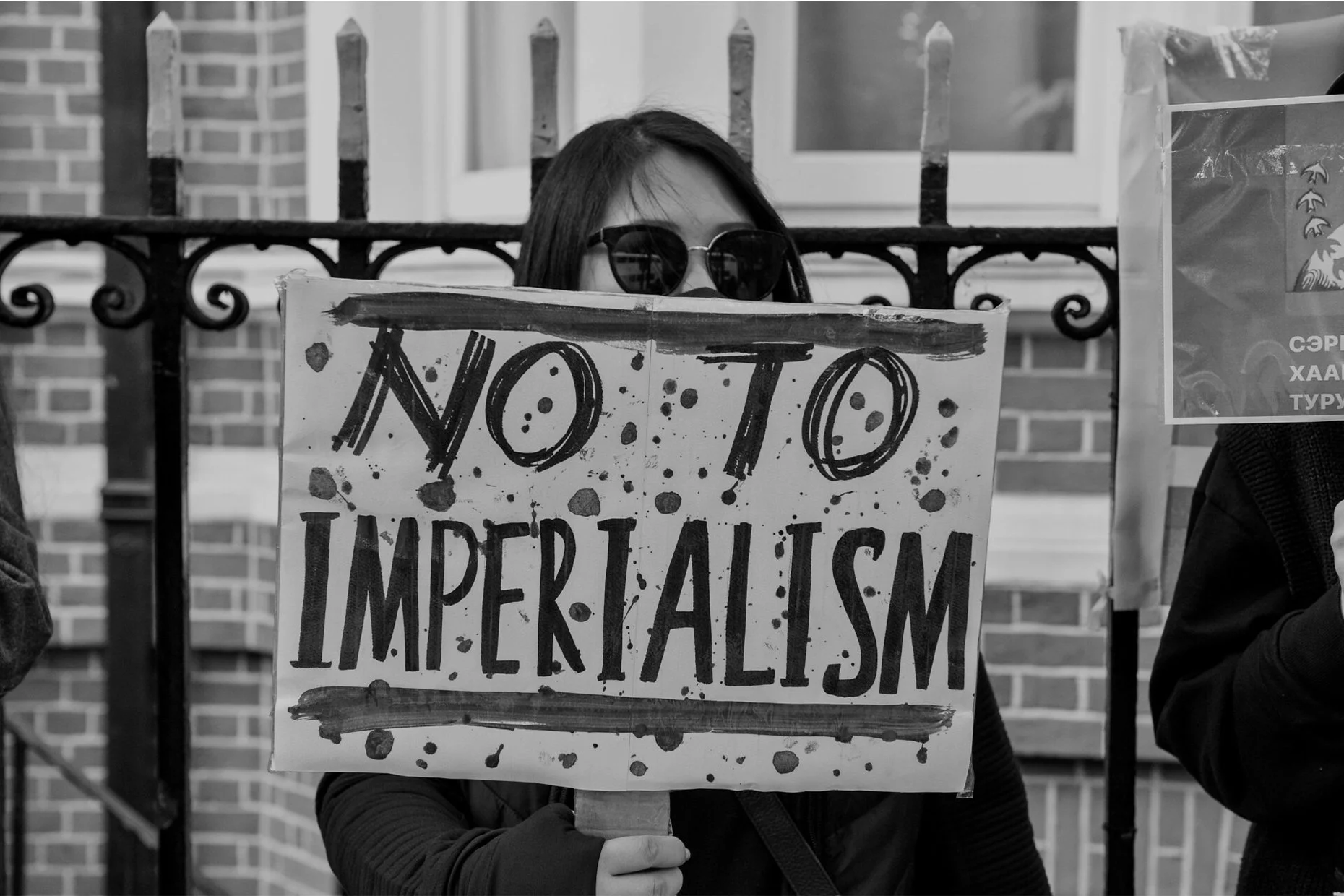A person wearing sunglasses holding a sign that says 'NO TO IMPERIALISM' during a protest, with a brick building and a fence behind them.