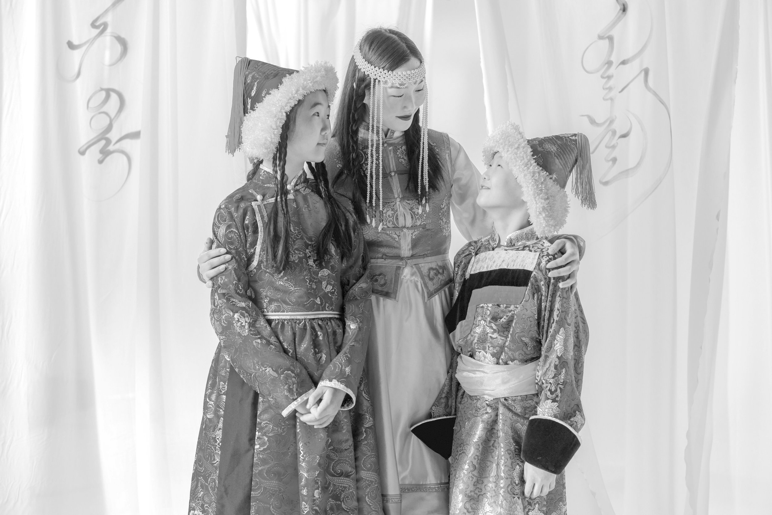 Three children dressed in traditional Asian costumes, with two girls wearing pointy hats, standing with a young woman also dressed in traditional attire, all facing each other indoors with a plain backdrop.
