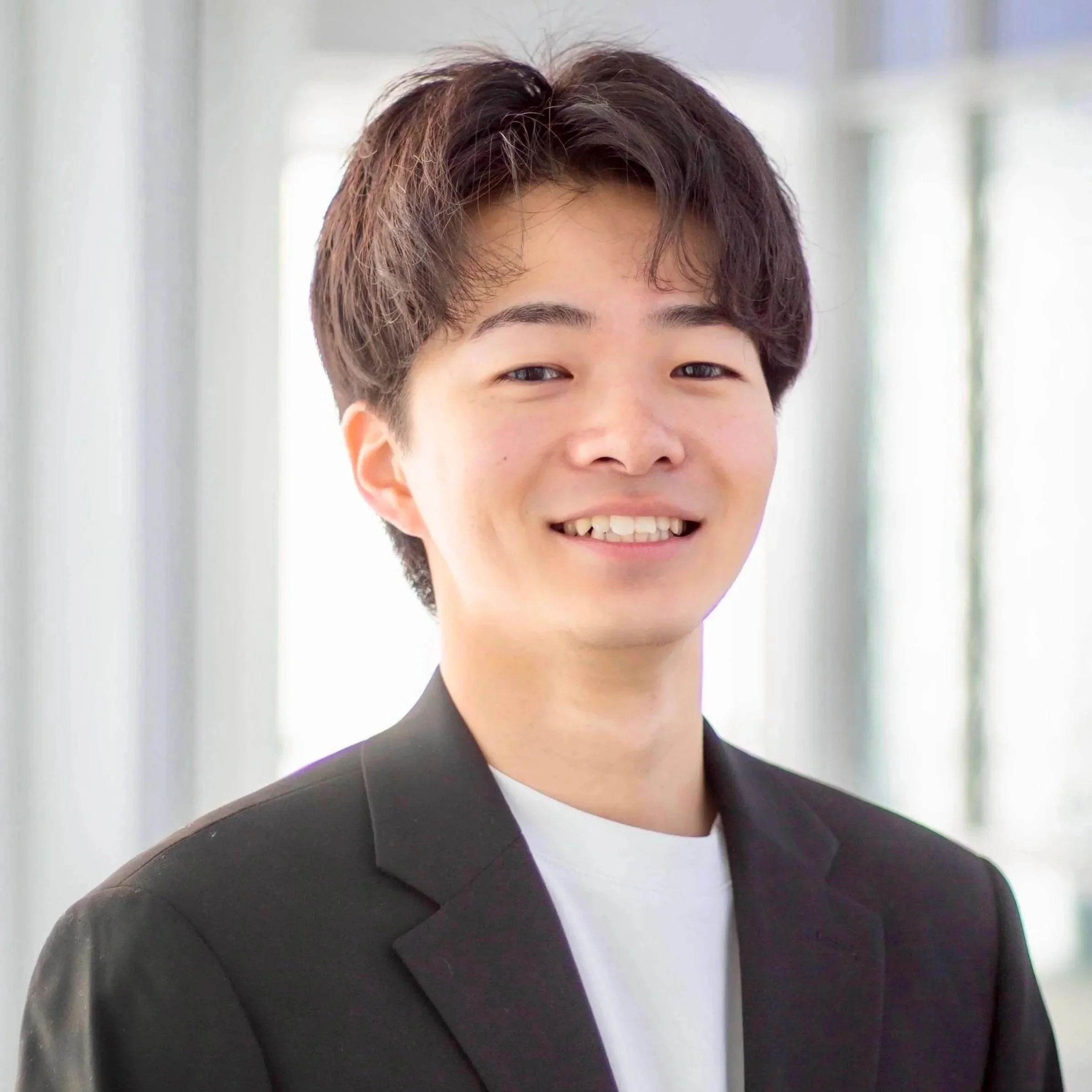 A young man with dark brown hair smiling, dressed in a black blazer and white shirt, in a bright, modern indoor setting.