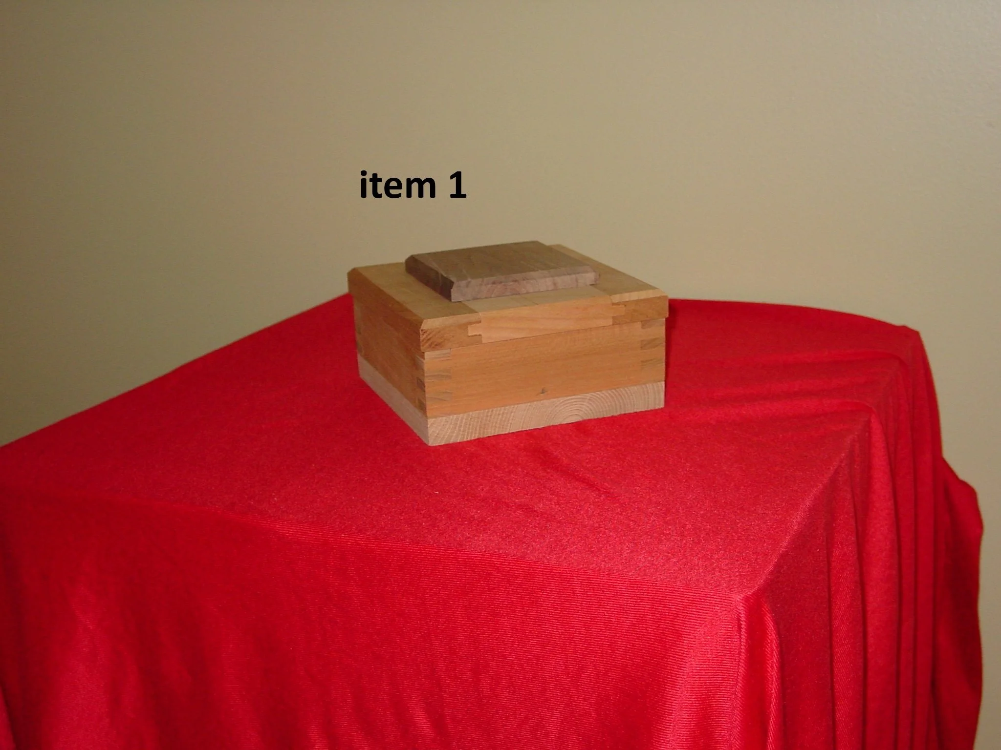 A wooden box with a smaller wooden piece on top, placed on a red draped surface against a beige wall. The box has a natural wood finish.