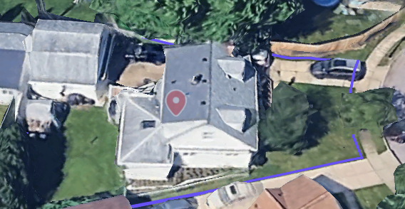 Aerial view of a house with a gray roof, surrounded by trees and a lawn, with a driveway and neighboring houses visible.